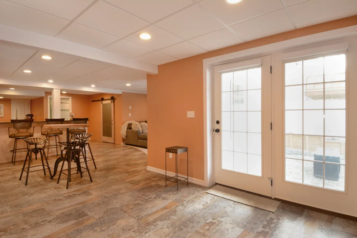 Spacious basement with peach walls, tiled floor, and recessed lighting. Features a bar area with stools, a sliding barn door, and French doors leading outside.