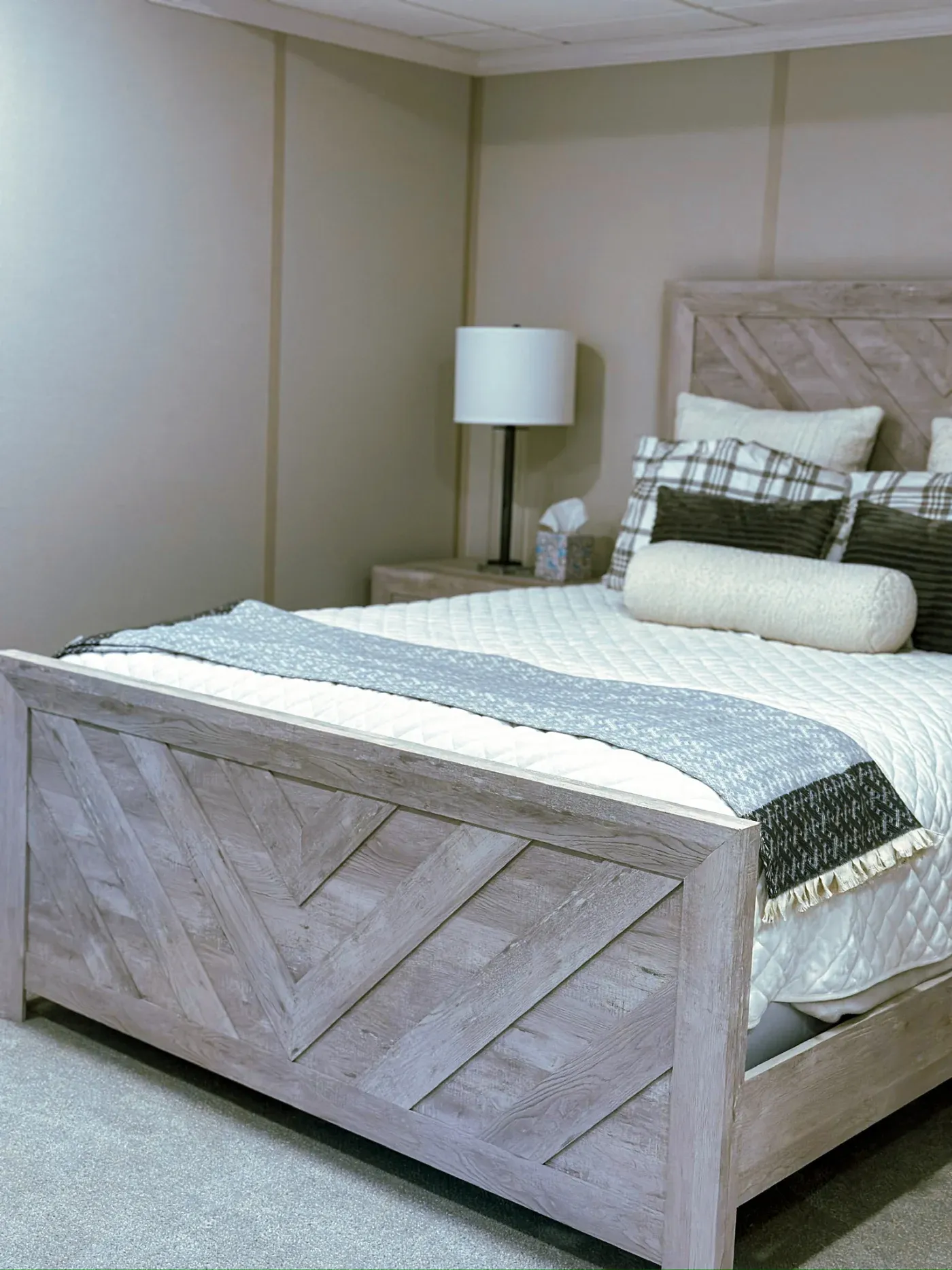 Minimalist bedroom featuring a grey wooden bed with geometric patterns, accented by plaid and textured pillows. Soft lighting creates a serene ambiance.