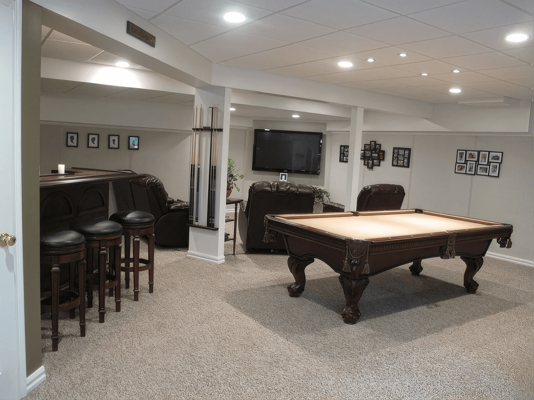 A cozy basement game room with a pool table, bar stools, a pool cue rack, leather recliners, a wall-mounted TV, and framed photos. Warm, inviting ambiance.