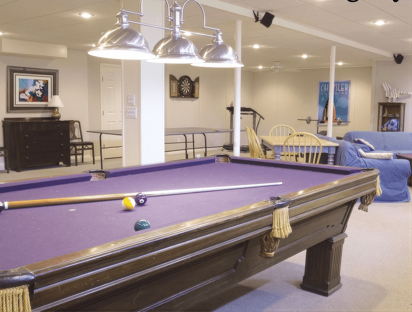 A cozy game room features a purple pool table with cue sticks, overhead lights, a dartboard, a blue sofa, wooden dining set, and playful decor.
