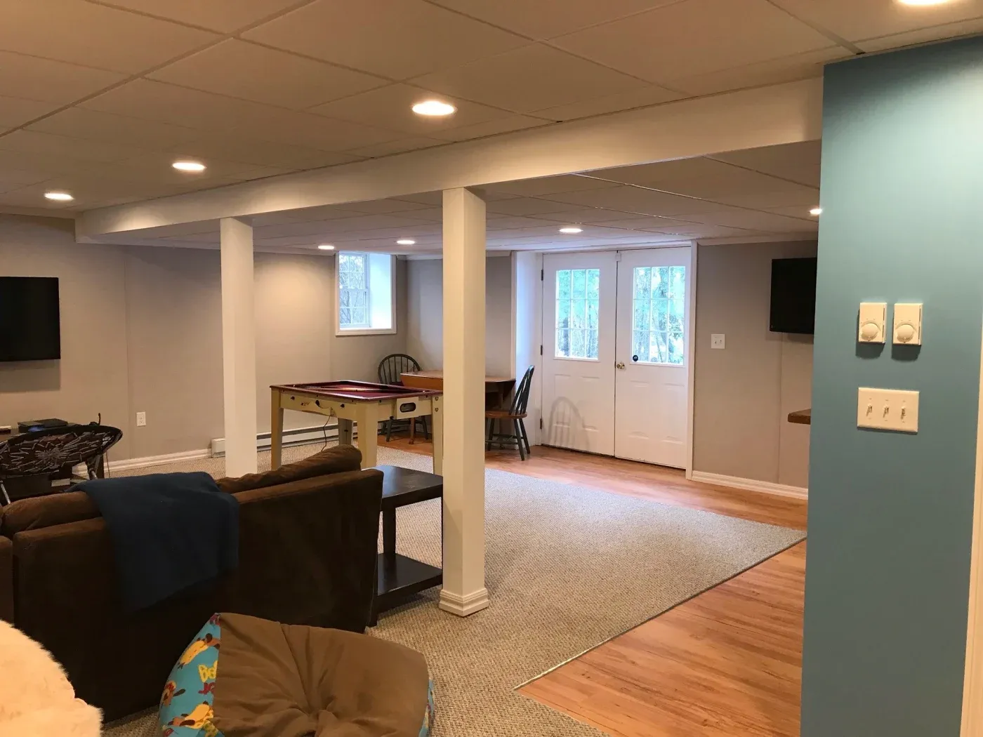 A cozy basement with beige walls and recessed lighting, featuring a brown couch, a small game table, and double doors leading outside.