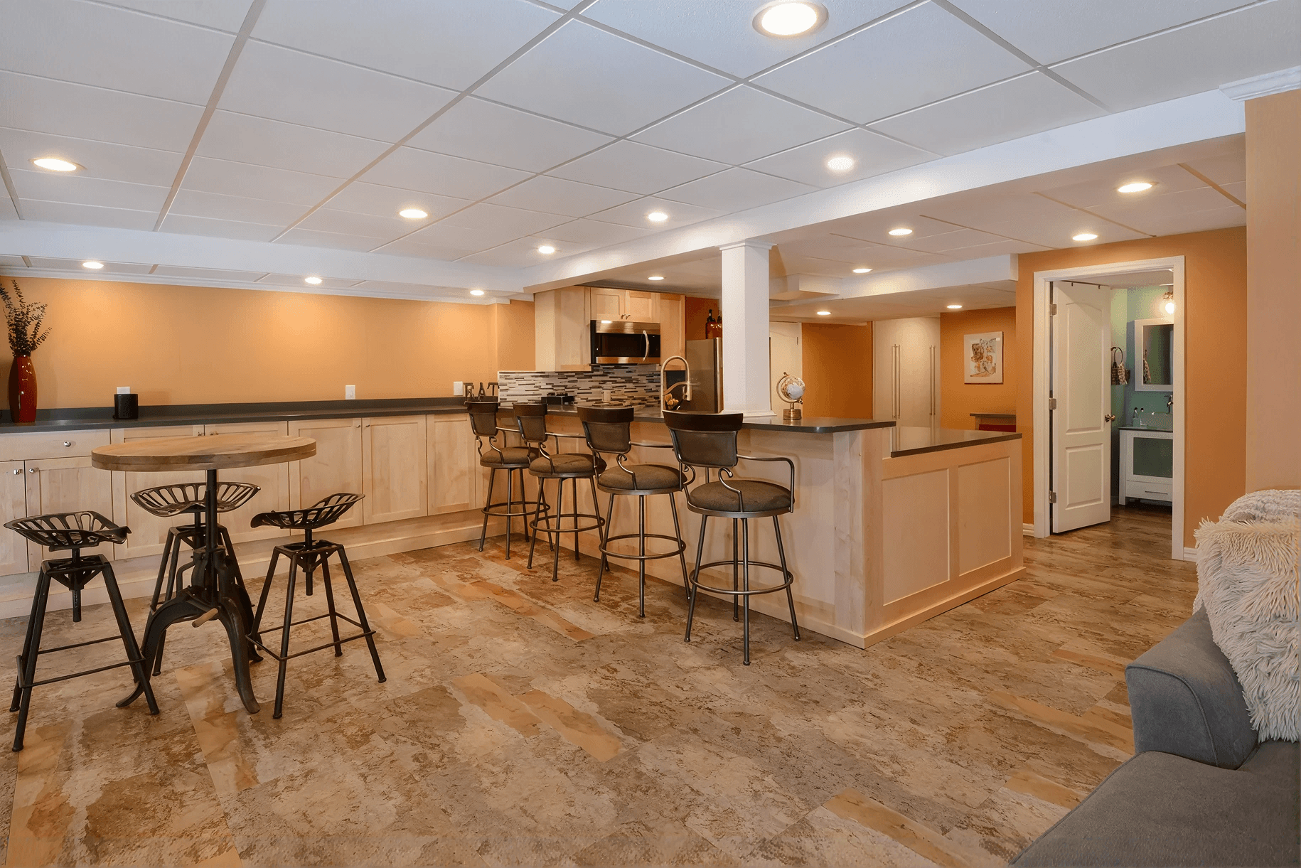 Spacious basement with rustic bar stools around a countertop, modern kitchen appliances, tan walls, and cozy seating, creating a warm, inviting atmosphere.
