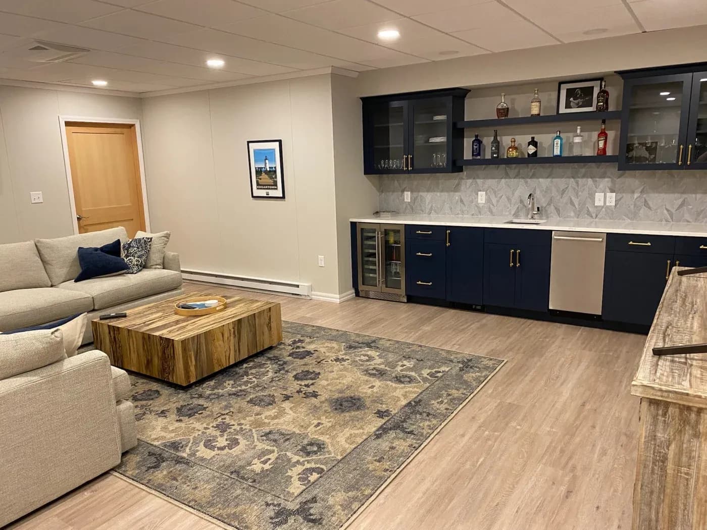 Cozy basement lounge with beige sectional sofa, blue and gray patterned rug, wooden coffee table, and a sleek bar area with navy cabinets and a mini fridge.
