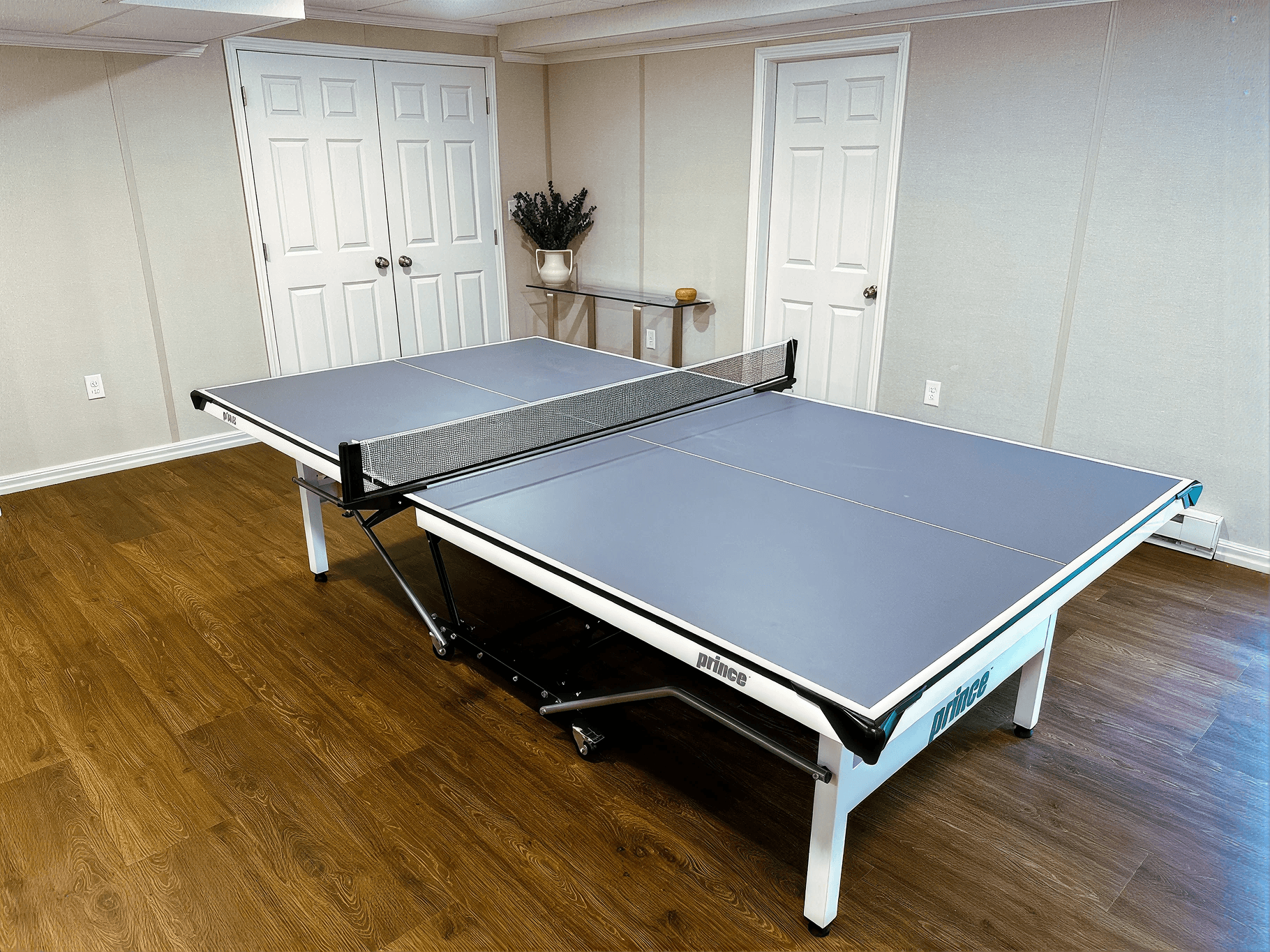 A ping pong table with a net is set up in a spacious, well-lit room with wooden floors and beige walls. Two closed white doors and a console table with a plant are in the background. The atmosphere is neat and ready for a game.