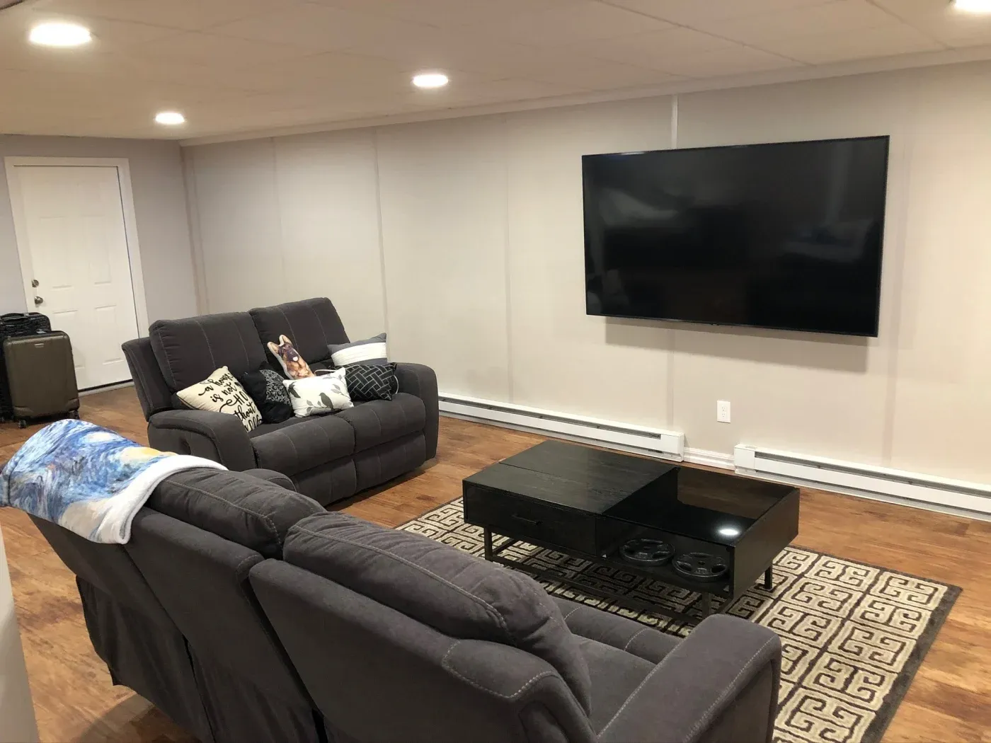 Modern living room with gray sofas, decorative pillows, and a blue throw. A large flat-screen TV hangs on the wall, and a black coffee table sits on a patterned rug.