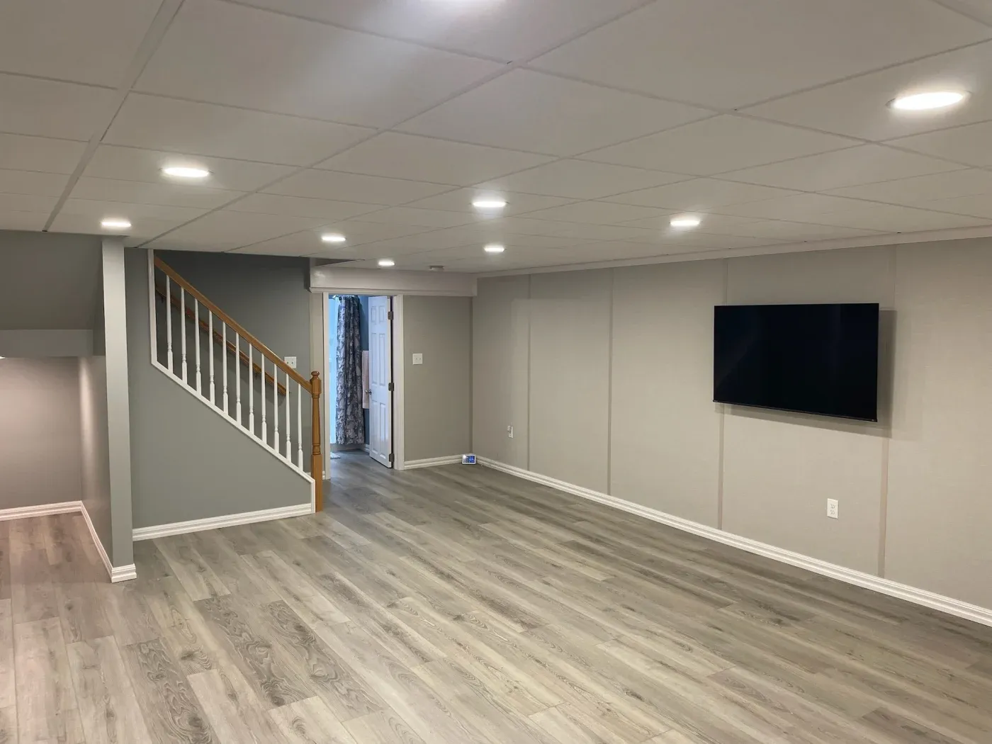Basement with gray walls and wooden flooring, featuring recessed lighting. A staircase with a wooden handrail and a wall-mounted television add modern touches.