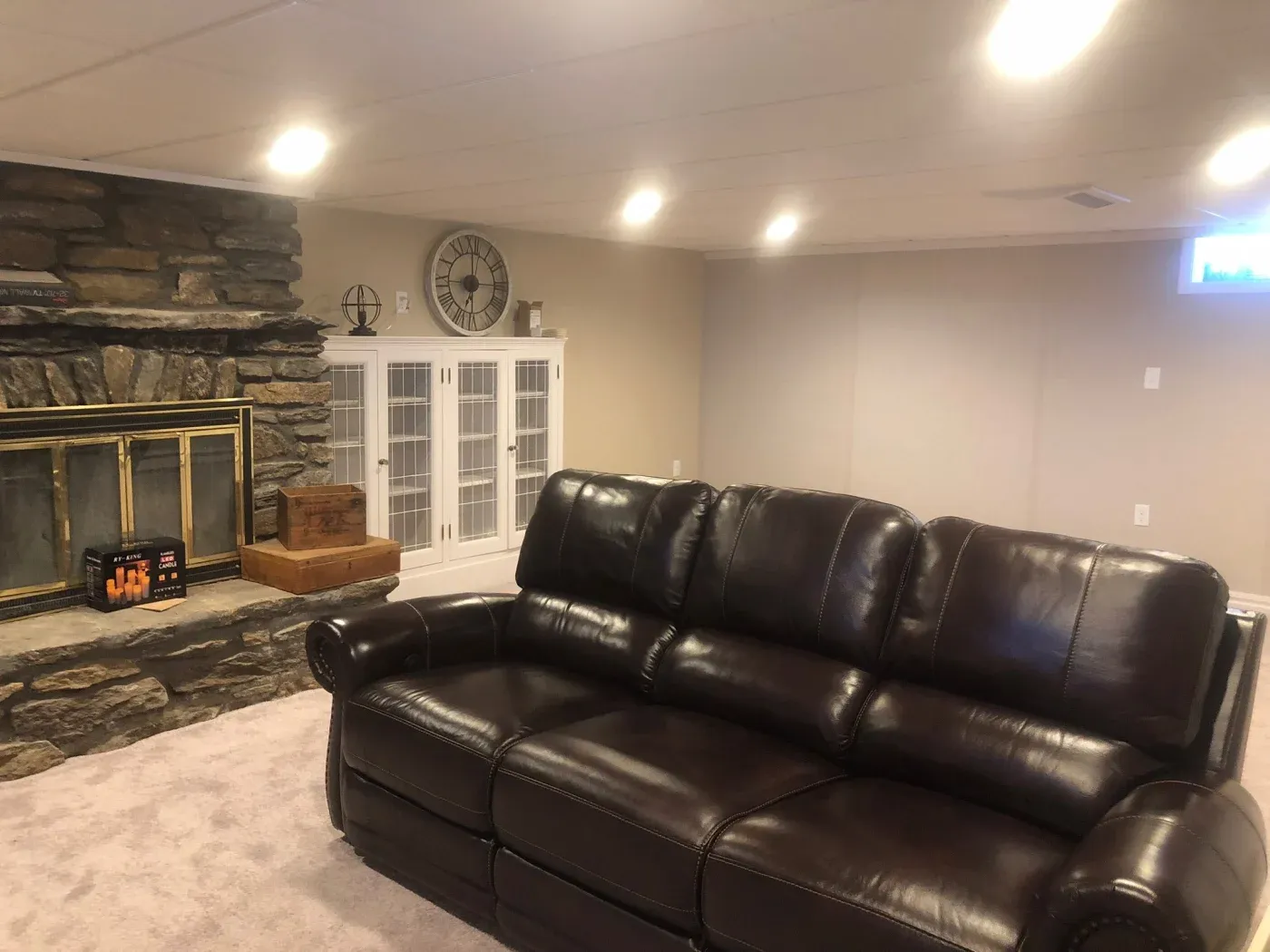 Basement living area with a dark brown leather sofa facing a stone fireplace. Soft lighting highlights a white cabinet and large wall clock. Cozy ambiance.