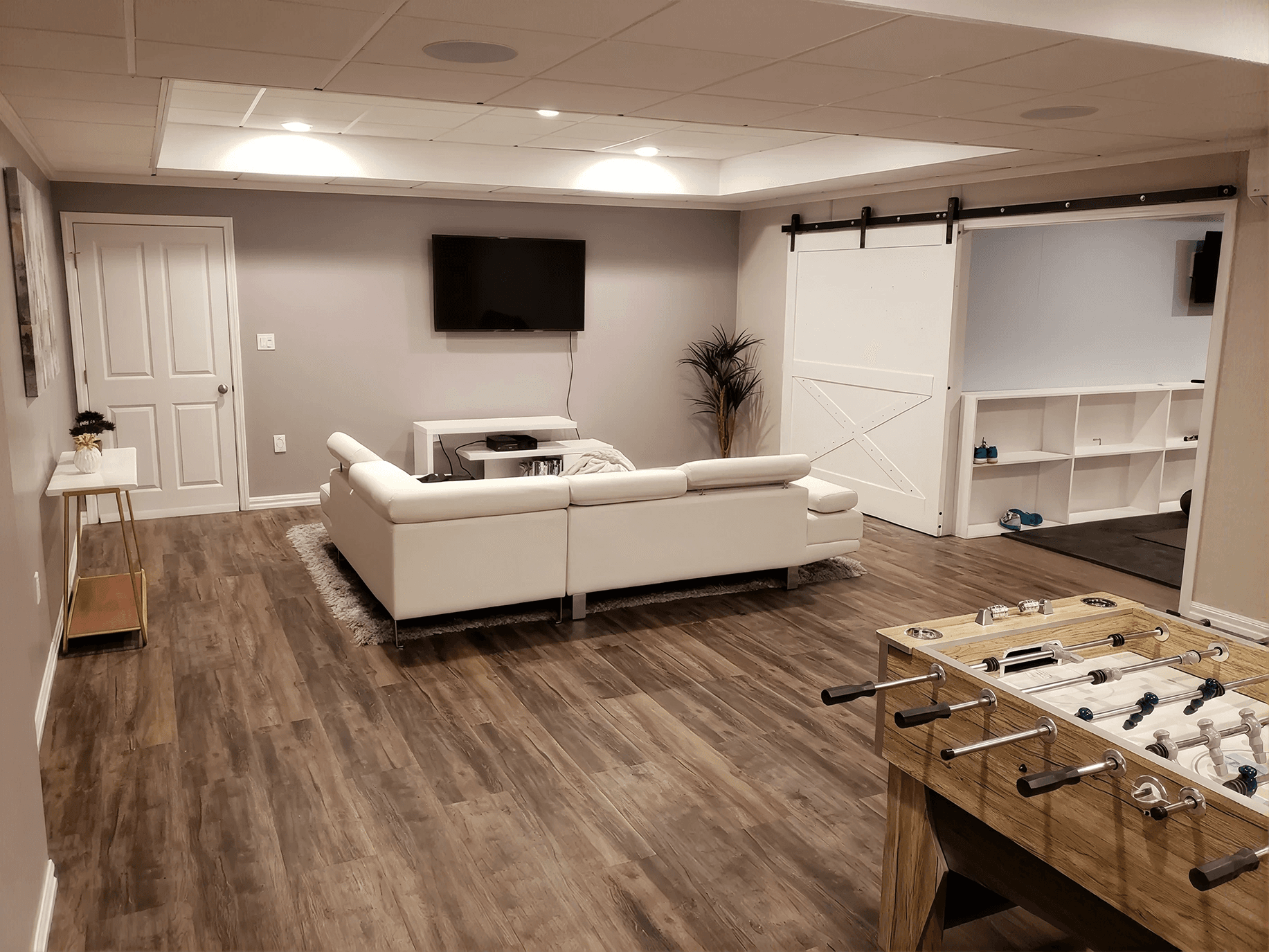 Modern basement with wood flooring, featuring a white sectional sofa facing a wall-mounted TV. Sliding barn door, foosball table, and indoor plant.