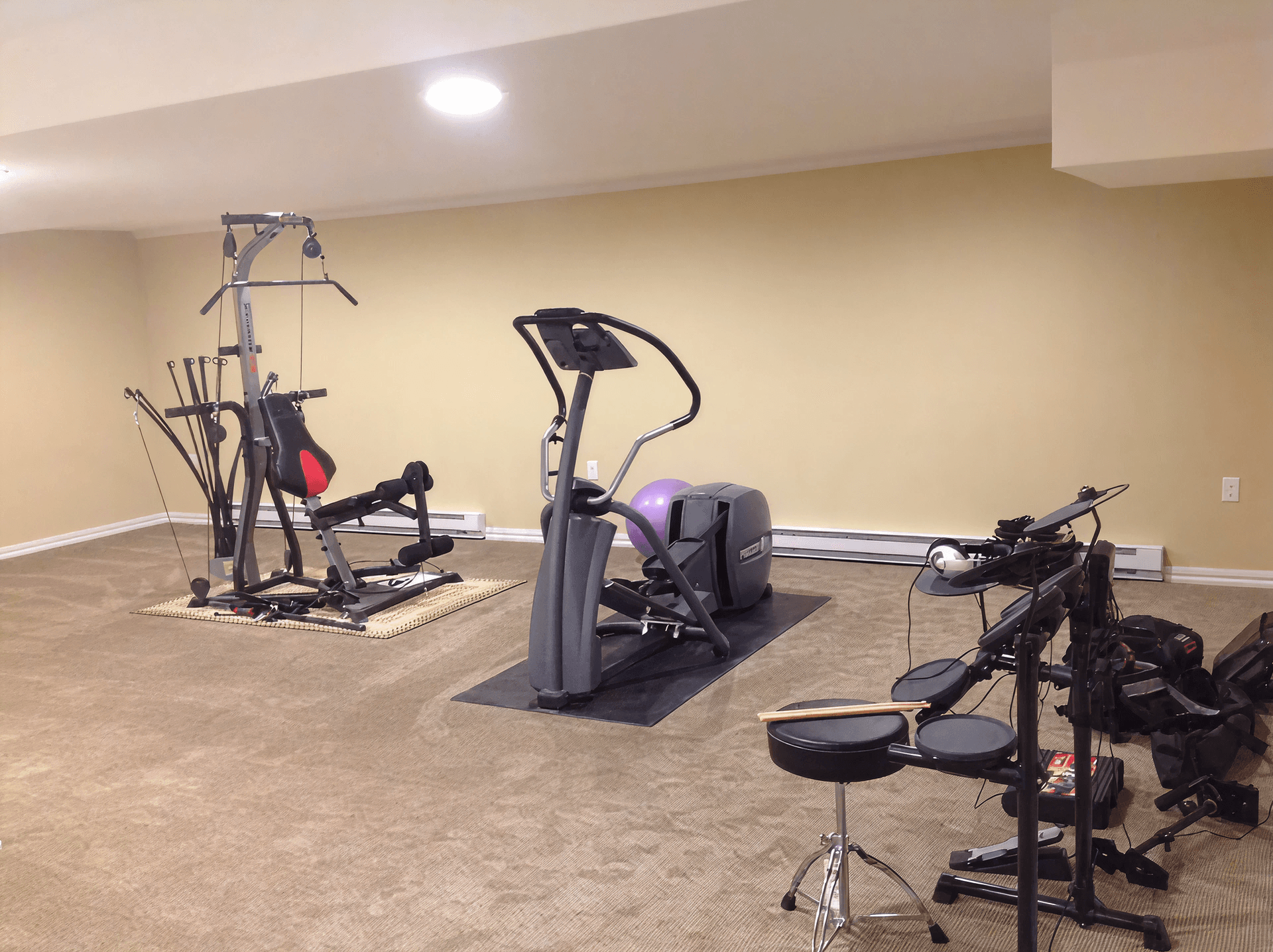 A home gym with beige walls and carpet features a multi-exercise machine, an elliptical, and an electronic drum kit. The space feels orderly and practical.
