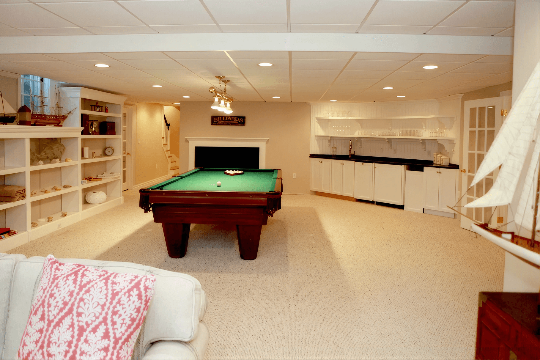 Bright basement entertainment room with a billiards table at the center, surrounded by white shelves with decor, and a cozy beige sofa with red patterned pillows.