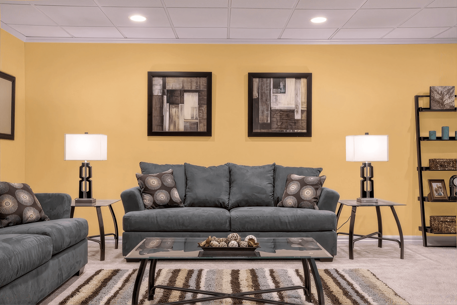A cozy living room with a gray sofa set against a warm yellow wall. Two abstract paintings hang above. Lamps on side tables emit soft light, complementing the rug's earthy tones.