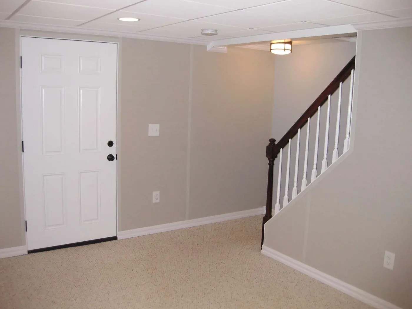 Finished basement recreation room with white six-panel door, carpeted flooring, beige walls, recessed lighting, and staircase with dark wood railing and white balusters