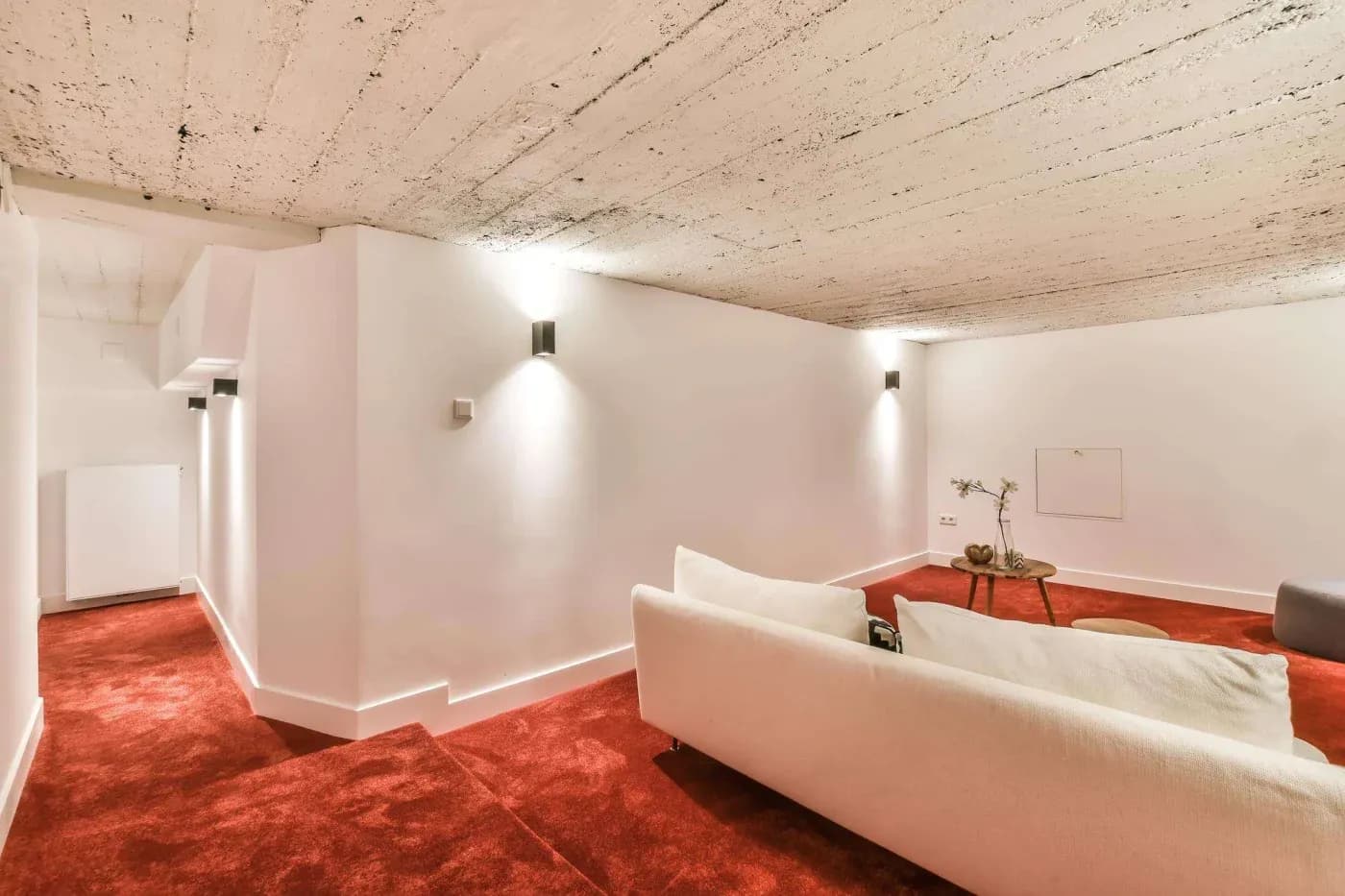 A minimalist room with white walls, a textured ceiling, red carpet, and a cream sofa. Soft lighting, a simple wooden table, and a peaceful ambiance.