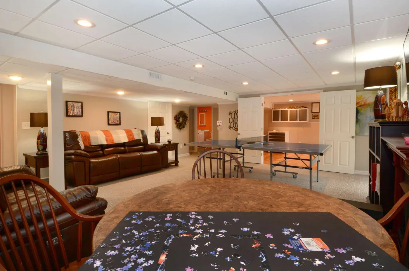 Spacious basement with a cozy ambiance featuring a leather sofa, decorative lamps, and wall art. A table with a puzzle sits nearby, and a ping pong table is in the back.