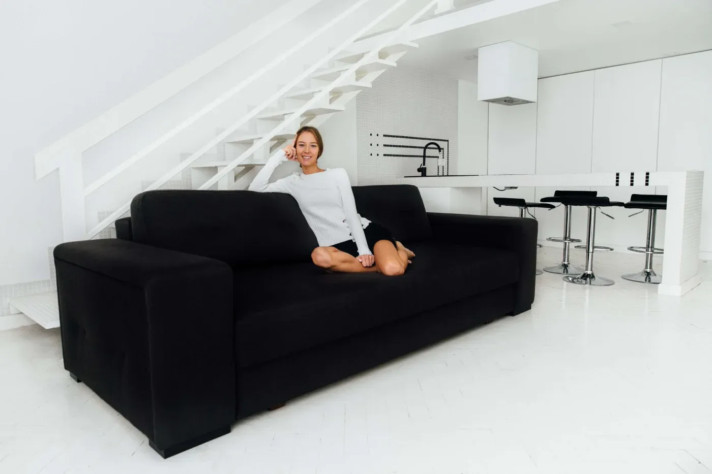 A woman in a white sweater sits on a black sofa in a modern, minimalist white room with a staircase and a sleek kitchen area visible in the background.
