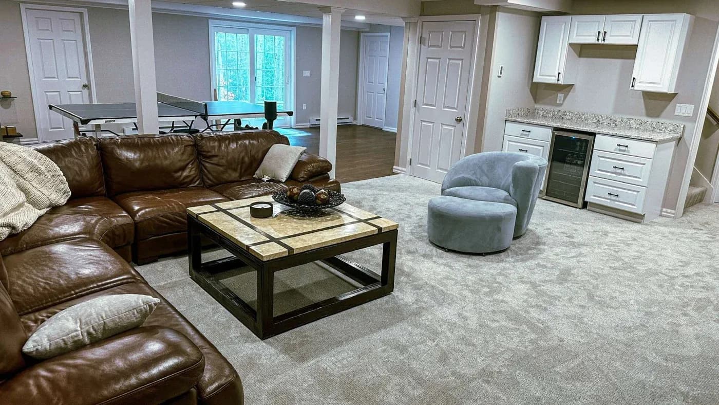 Spacious basement with a plush brown leather sectional, soft gray armchair, and elegant marble coffee table. Nearby, a ping pong table adds a playful touch.