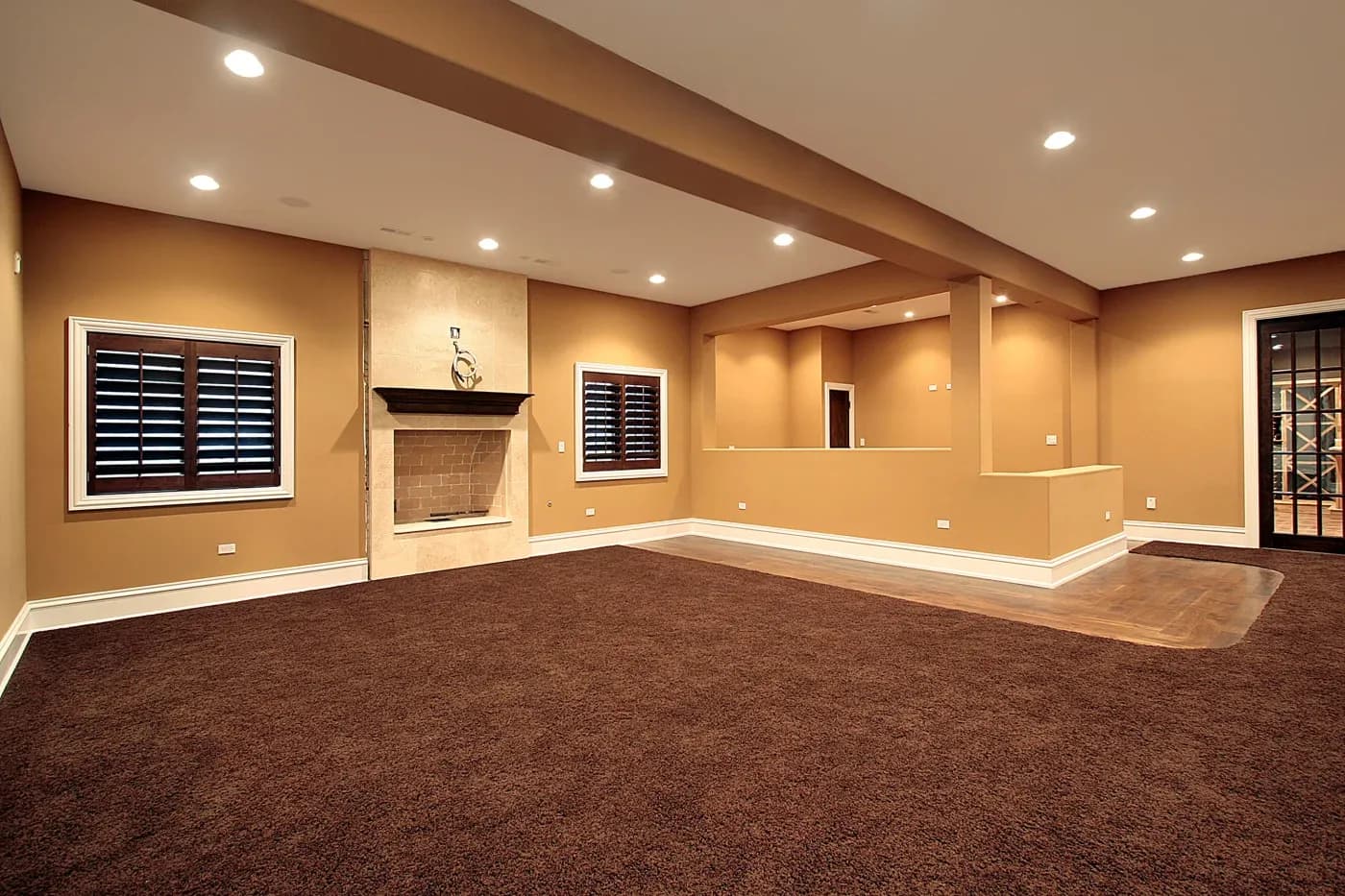 Spacious basement with warm beige walls, dark brown carpet, recessed lighting, fireplace, and big windows with dark shutters. Cozy and inviting atmosphere.