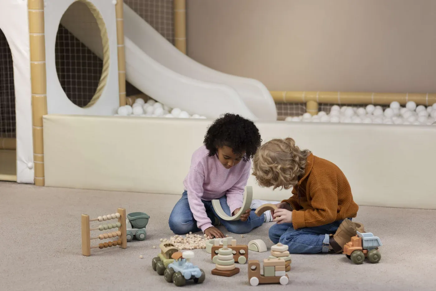 Two children play with wooden toys on carpet in a playroom, near a slide and ball pit. The scene conveys a sense of fun and creativity.