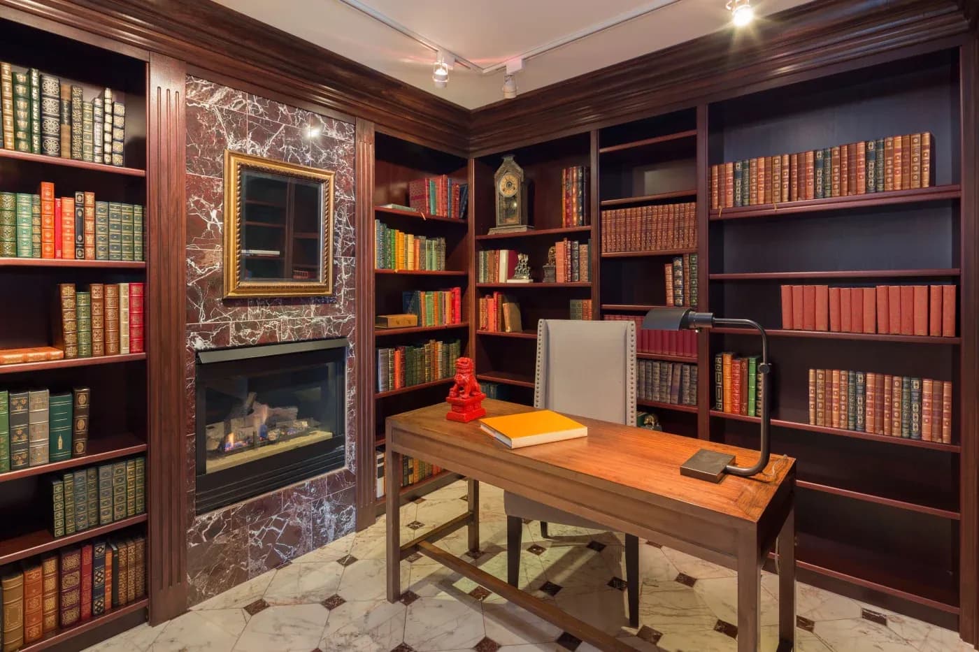 Cozy library with wooden shelves filled with colorful books, a marble fireplace, and a wooden desk with a chair, conveying warmth and elegance.