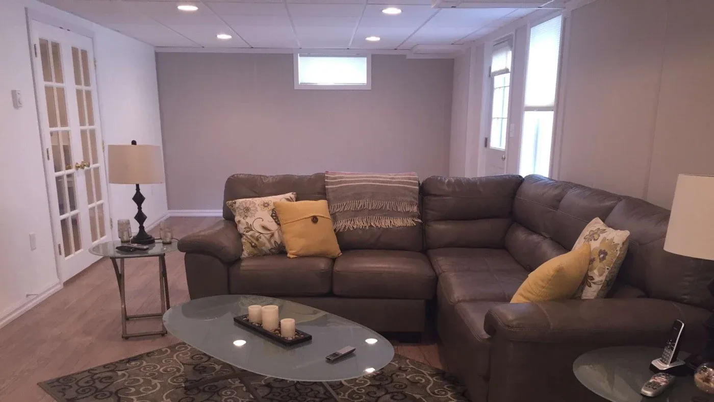 A cozy living room with a large brown sectional sofa adorned with yellow and patterned pillows. A glass coffee table with candles is in front, flanked by two lamps. The room has soft lighting and French doors, creating a warm and inviting ambiance.