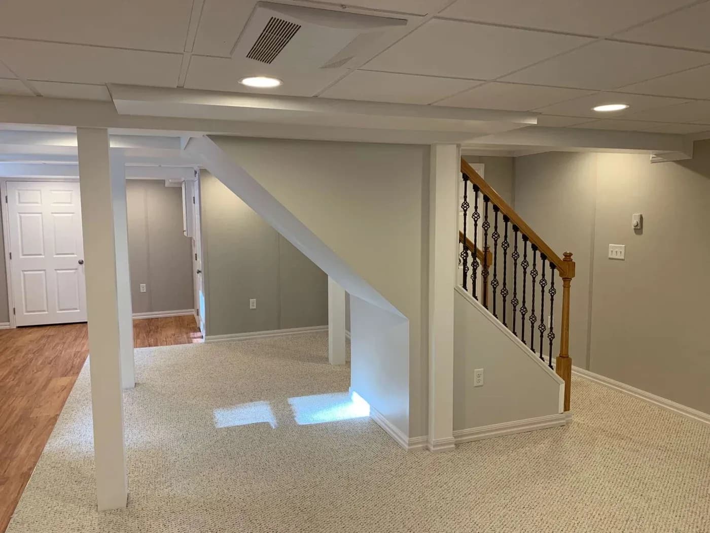 A modern basement with beige walls, white pillars, carpeted and wood floors, and a staircase with wooden handrail and black balusters. Bright and inviting.