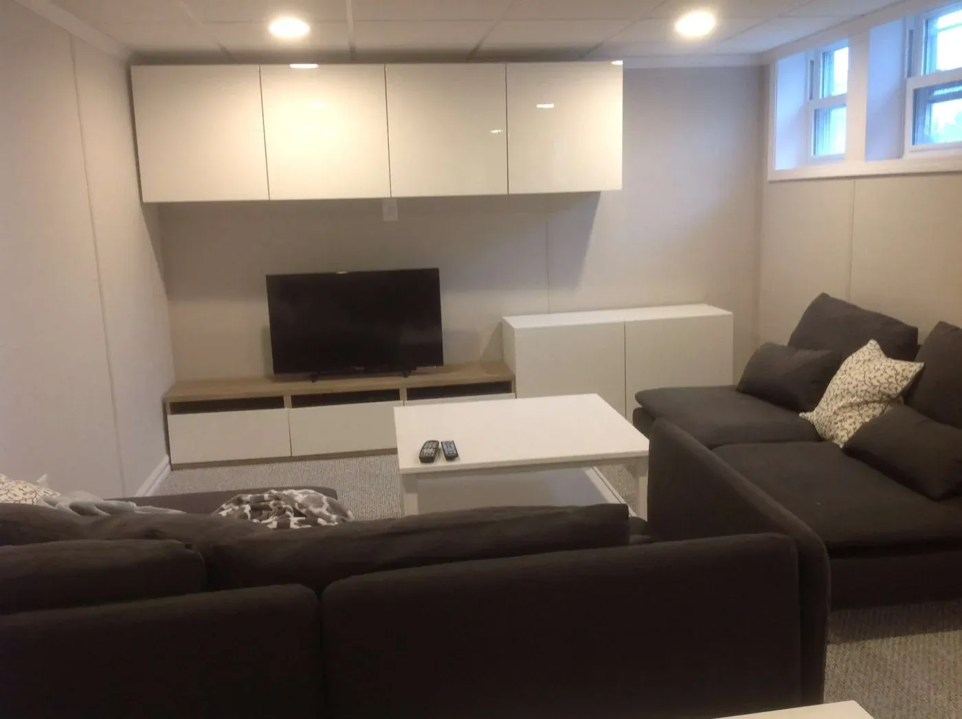 A cozy living room with a modern design features a dark sectional couch and patterned pillows. A flat-screen TV sits on a low console opposite a white coffee table with remote controls. Bright, soft lighting and minimalistic wall cabinets enhance the room’s inviting tone.