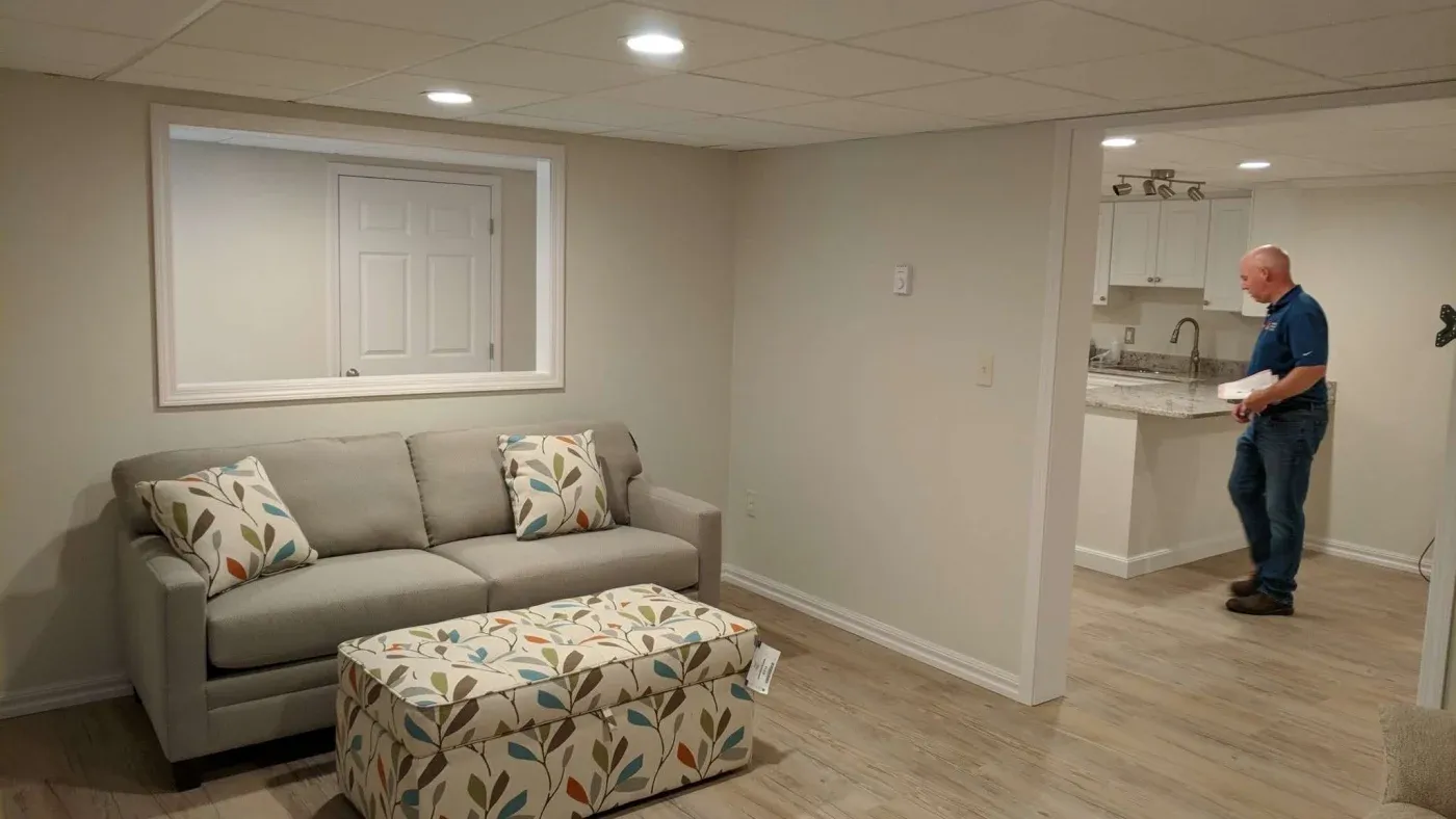 A cozy basement living area with light gray walls. A gray sofa with leaf-patterned pillows faces a matching patterned ottoman. A person stands in the adjacent kitchenette.