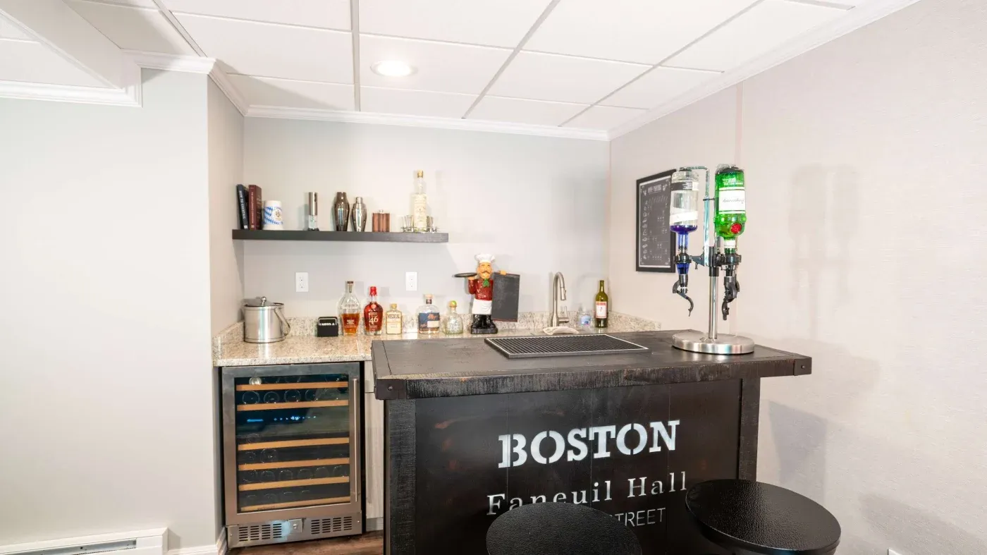 Cozy home bar setup with a granite countertop and dark wood finish. Bottles and glassware on shelves, drink dispensers on the counter. Modern, inviting feel.