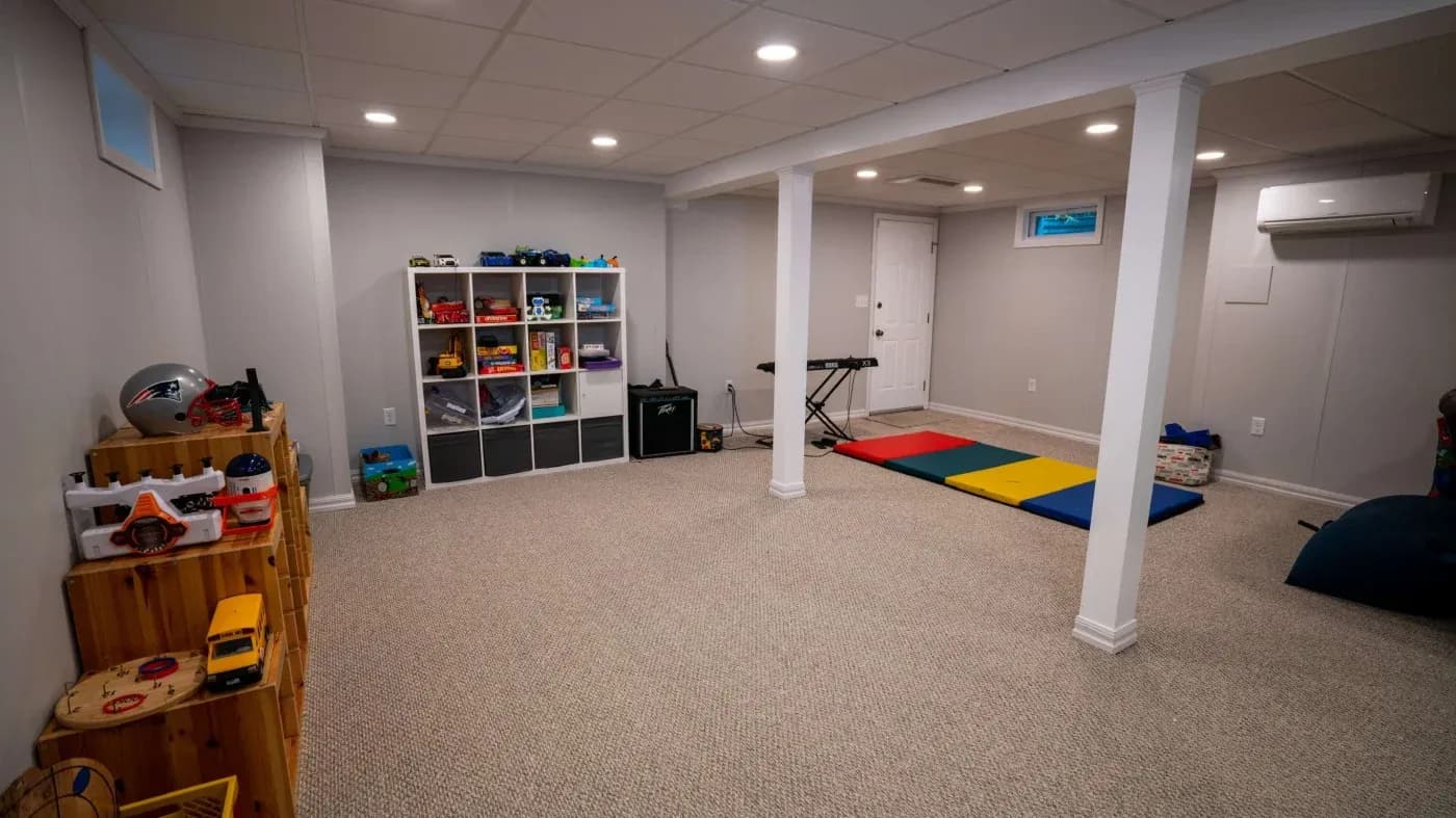 A spacious, neatly organized playroom with carpeted floor, white walls, and ceiling lights. Shelves hold colorful toys, while mats and a keyboard lay nearby.