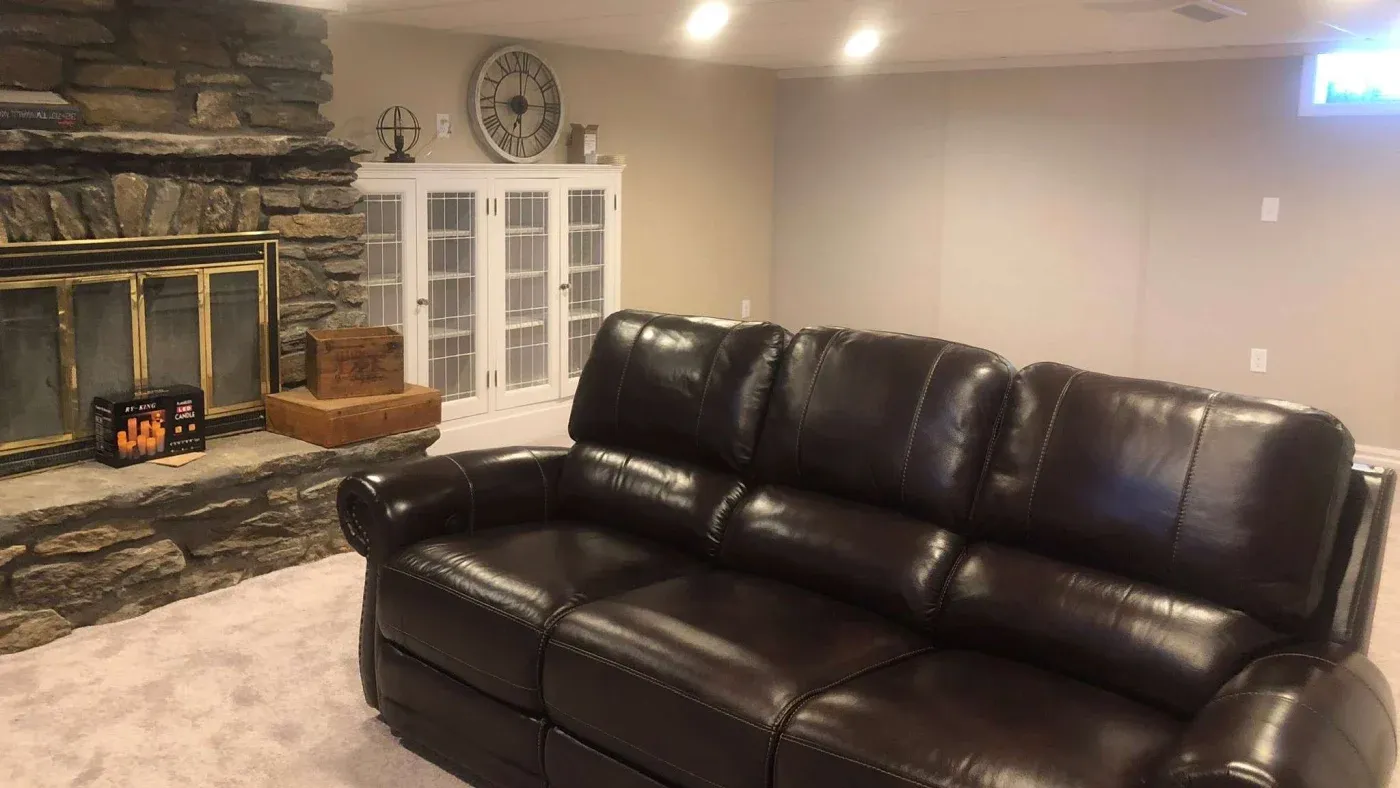 A cozy living room features a dark leather sofa, a stone fireplace with a brass screen, and a white cabinet with glass doors. The atmosphere is warm and inviting.