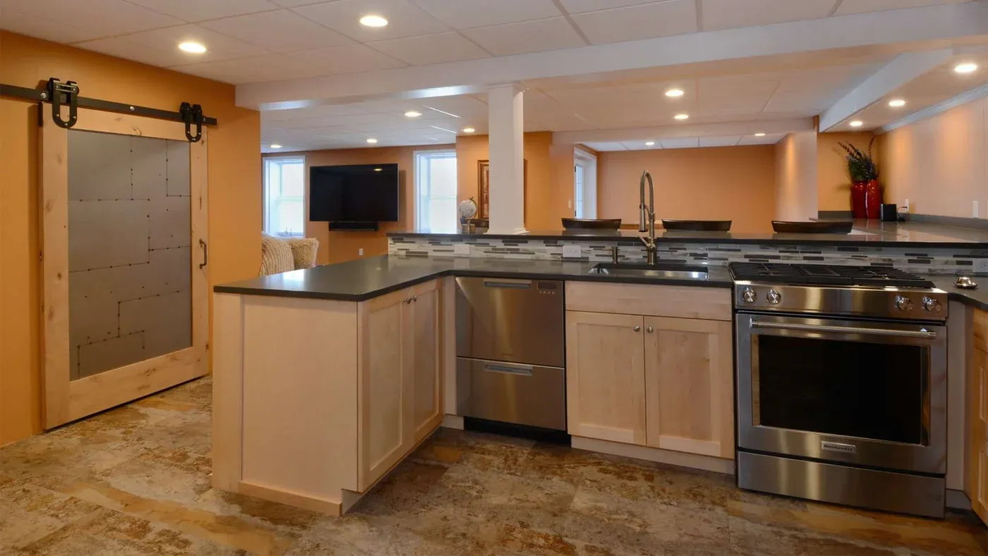 A modern kitchen with warm orange walls features light wood cabinets, stainless steel appliances, and a black countertop. A sliding barn door adds rustic charm.
