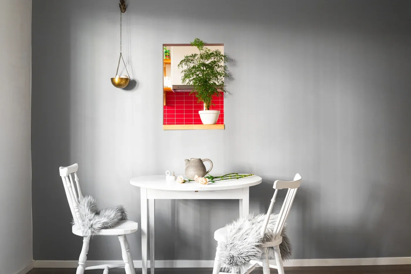 Cozy dining area with two white chairs, a round table, and soft gray cushions. A mirror reflects a plant and red tiles, adding color to the gray wall.