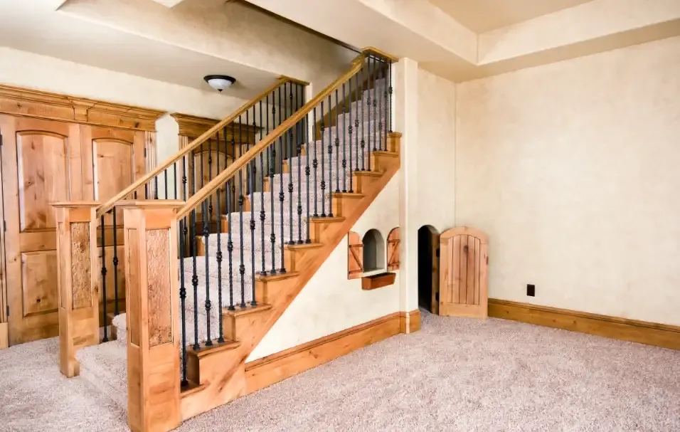 A carpeted staircase with wooden railings and dark metal balusters leads to a basement. Nearby, a small door under the stairs adds a whimsical touch.