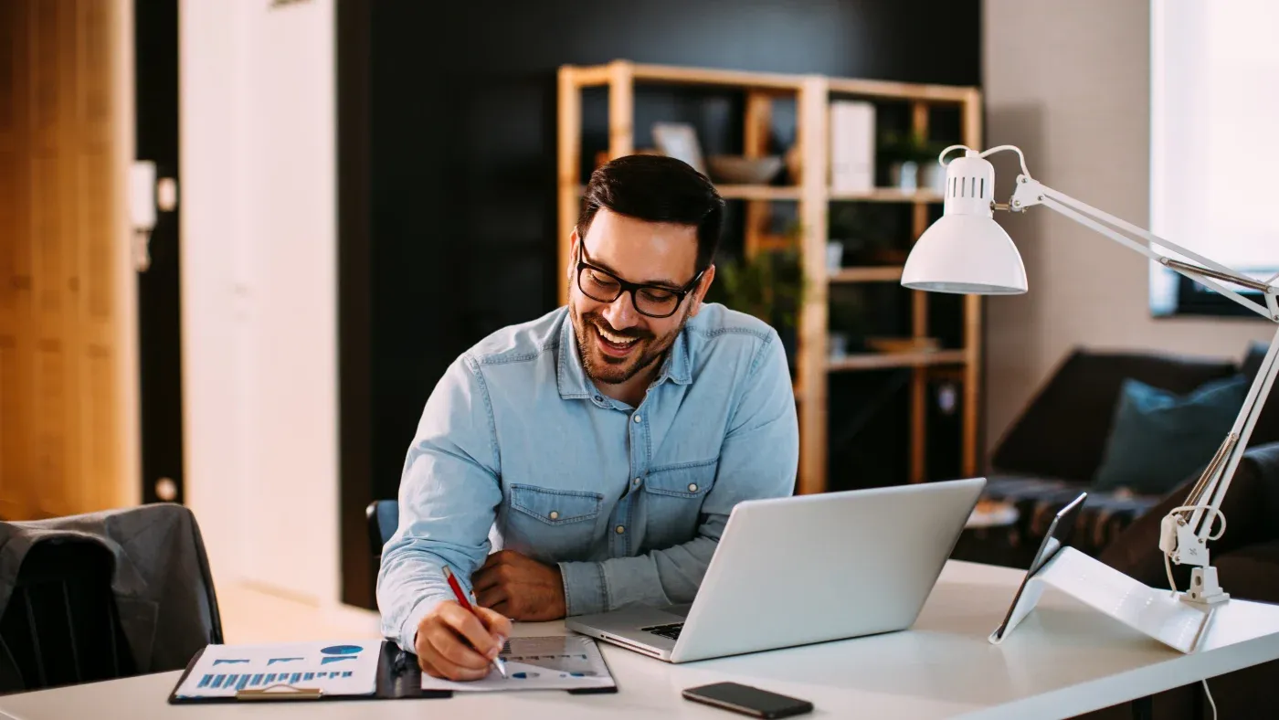 A man in glasses, wearing a blue shirt, smiles while working at a desk with a laptop, documents, and a lamp. The setting conveys a productive, positive atmosphere.