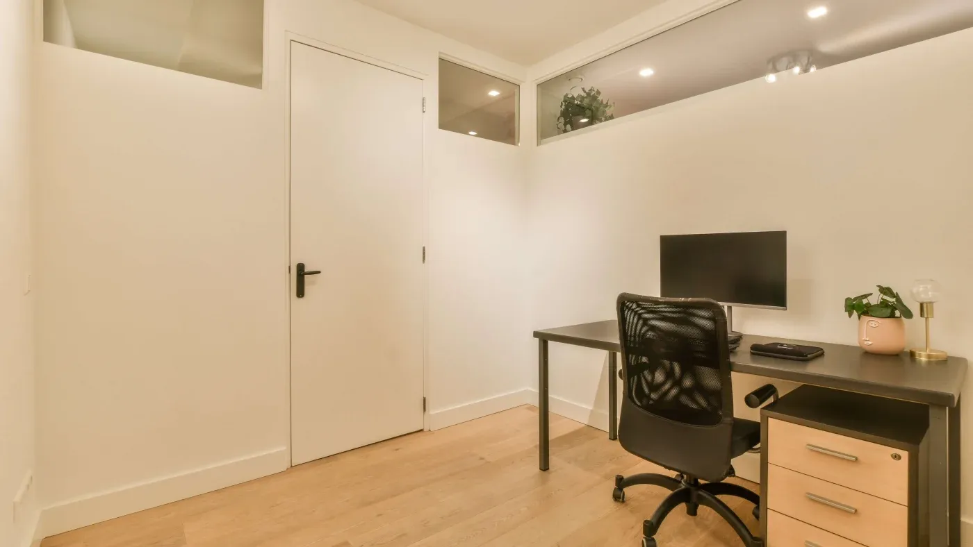 A minimalistic home office with a sleek black desk, monitor, and black mesh chair. Light wood flooring, white walls, and potted plant create a calm atmosphere.