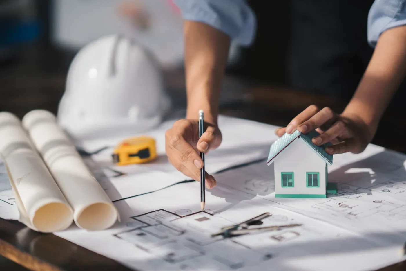 Close-up of hands working on architectural blueprints with a pencil and a small house model. Nearby are rolled-up plans, a white hard hat, and a tape measure, conveying a theme of construction planning.