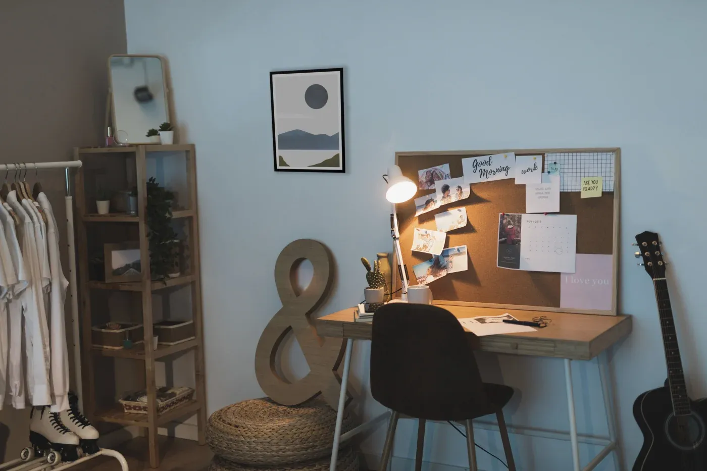 Cozy workspace with a wooden desk, bulletin board, and lamp. Clothes rack, guitar, and plants add personality. Soft lighting creates a calm ambiance.