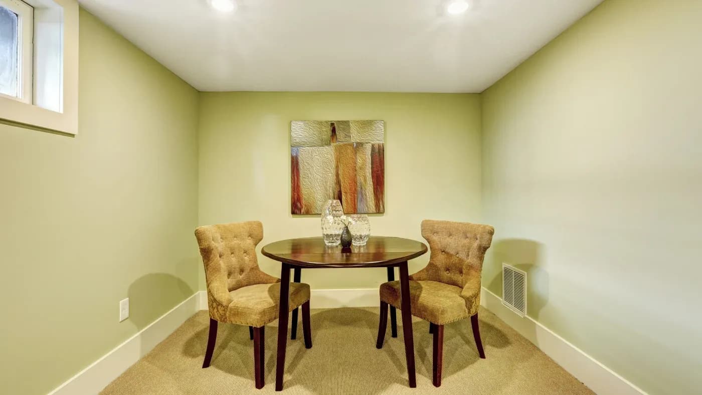 A cozy room with light green walls features a round wooden table and two beige upholstered chairs. An abstract painting hangs on the wall above.