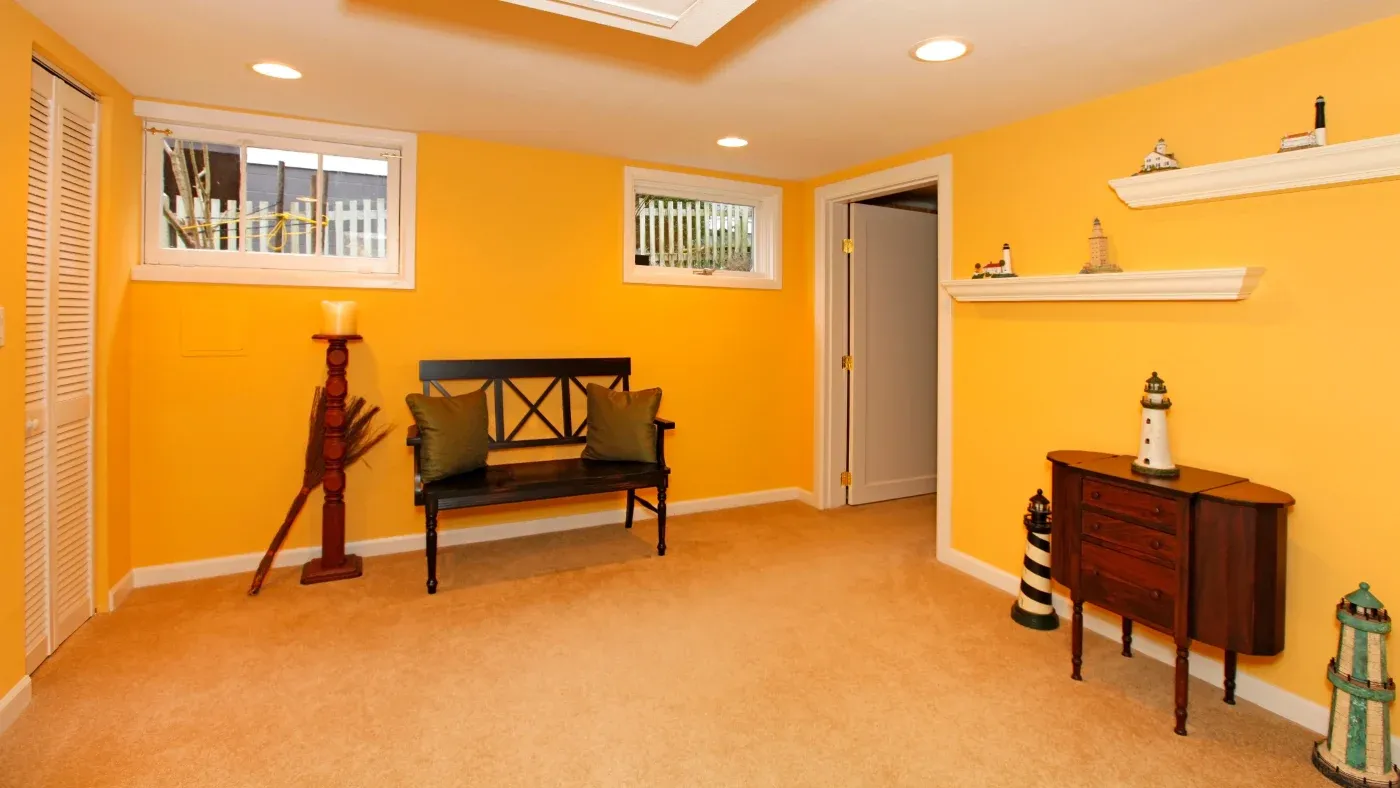 A warm, cozy basement with bright yellow walls and beige carpet features a wooden bench with green cushions, nautical-themed decor, and a small cabinet.