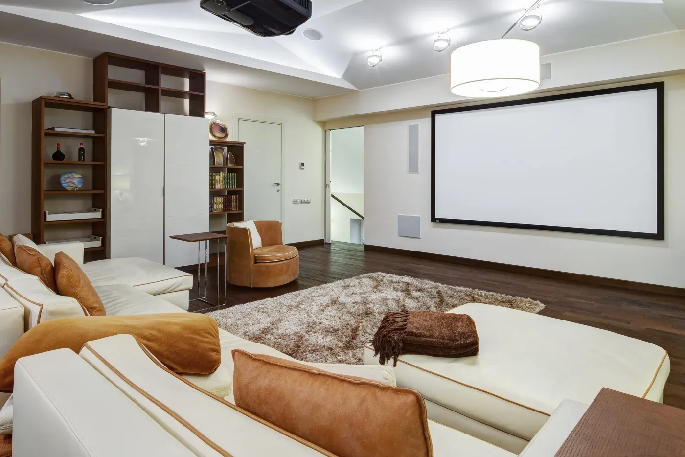 Cozy home theater with white sofas, tan cushions, a large projector screen, bookshelf, and a plush rug, creating a warm and inviting ambiance.
