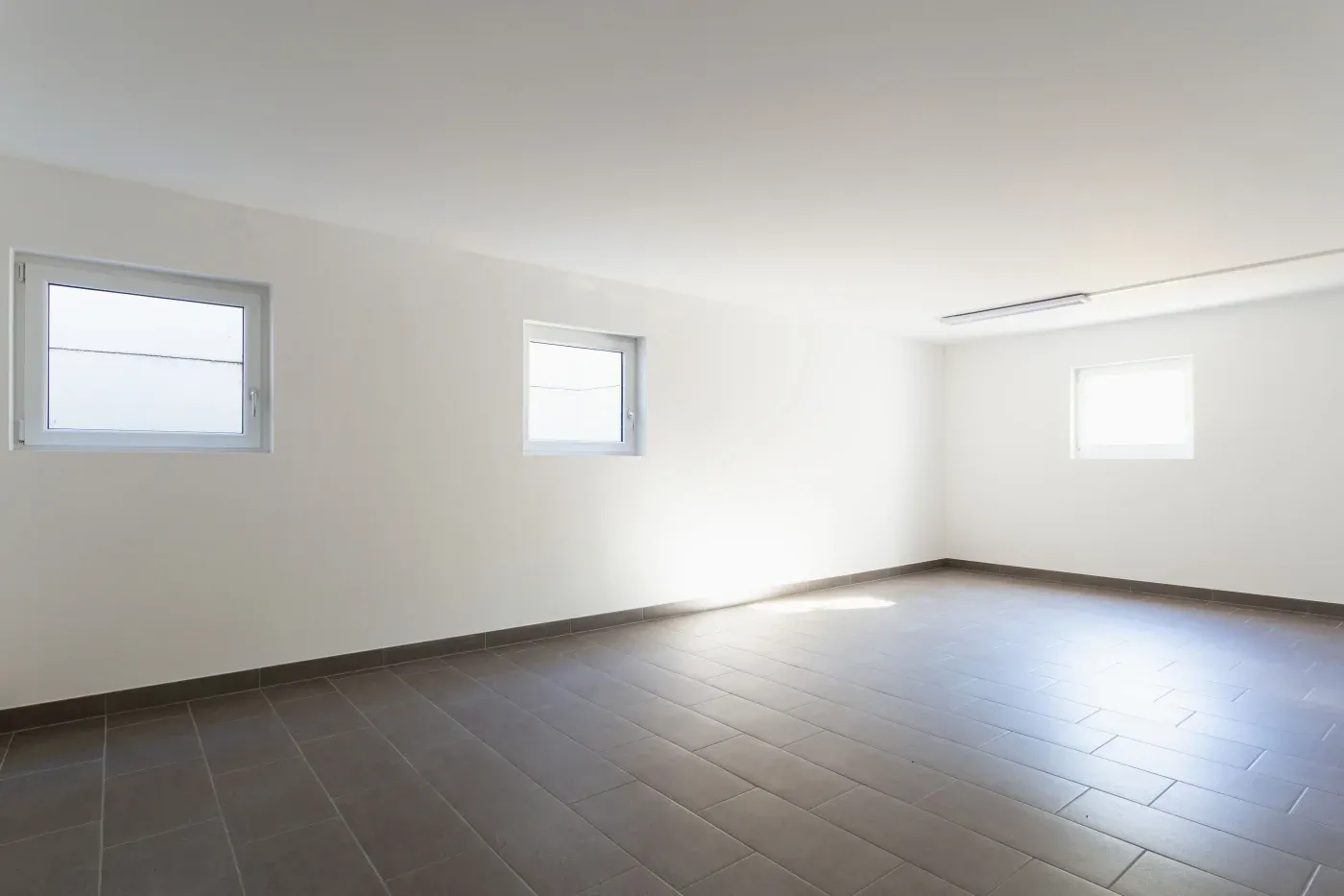 Bright, empty room with three small windows, white walls, and gray tiled flooring. Sunlight streams in, creating a clean and minimalist atmosphere.