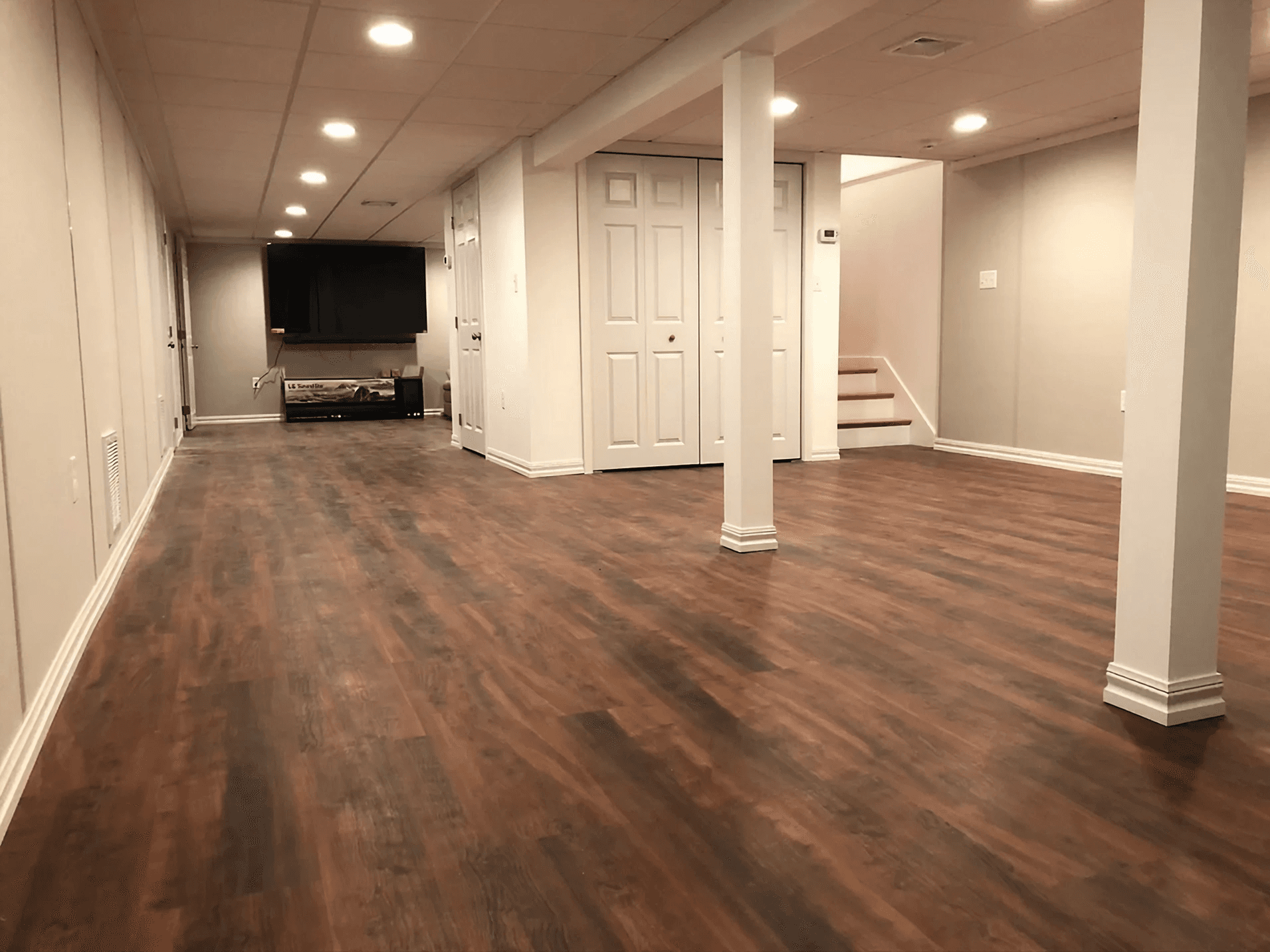 A spacious basement with polished wood flooring, white walls, and a recessed ceiling. It features a flat-screen TV, a closet, and minimal furnishings, creating a modern, open atmosphere. Spotlights illuminate the area warmly.