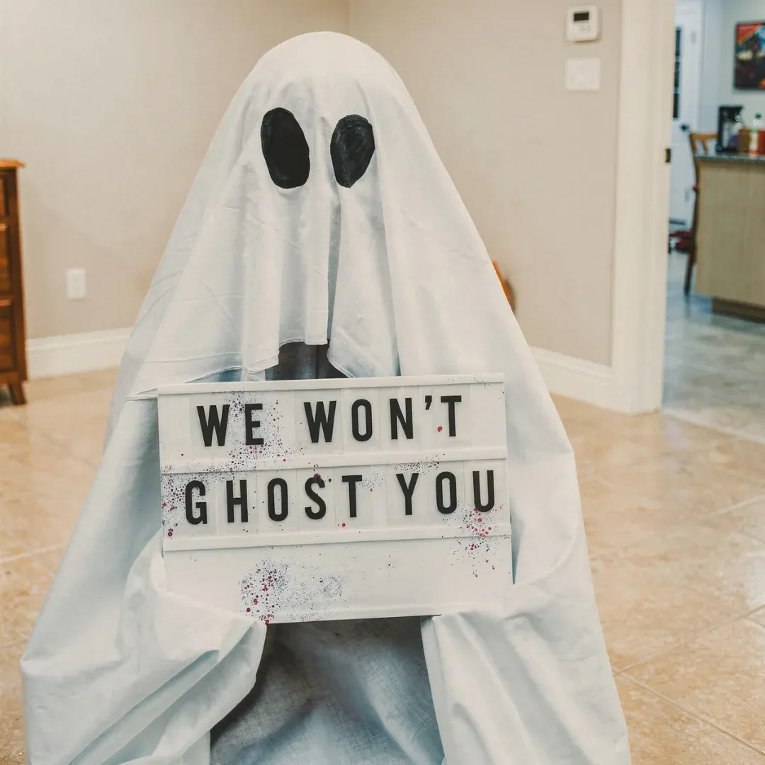A person dressed as a ghost in a white sheet with black eye holes holds a sign reading "We Won't Ghost You" in a lighthearted, playful setting.
