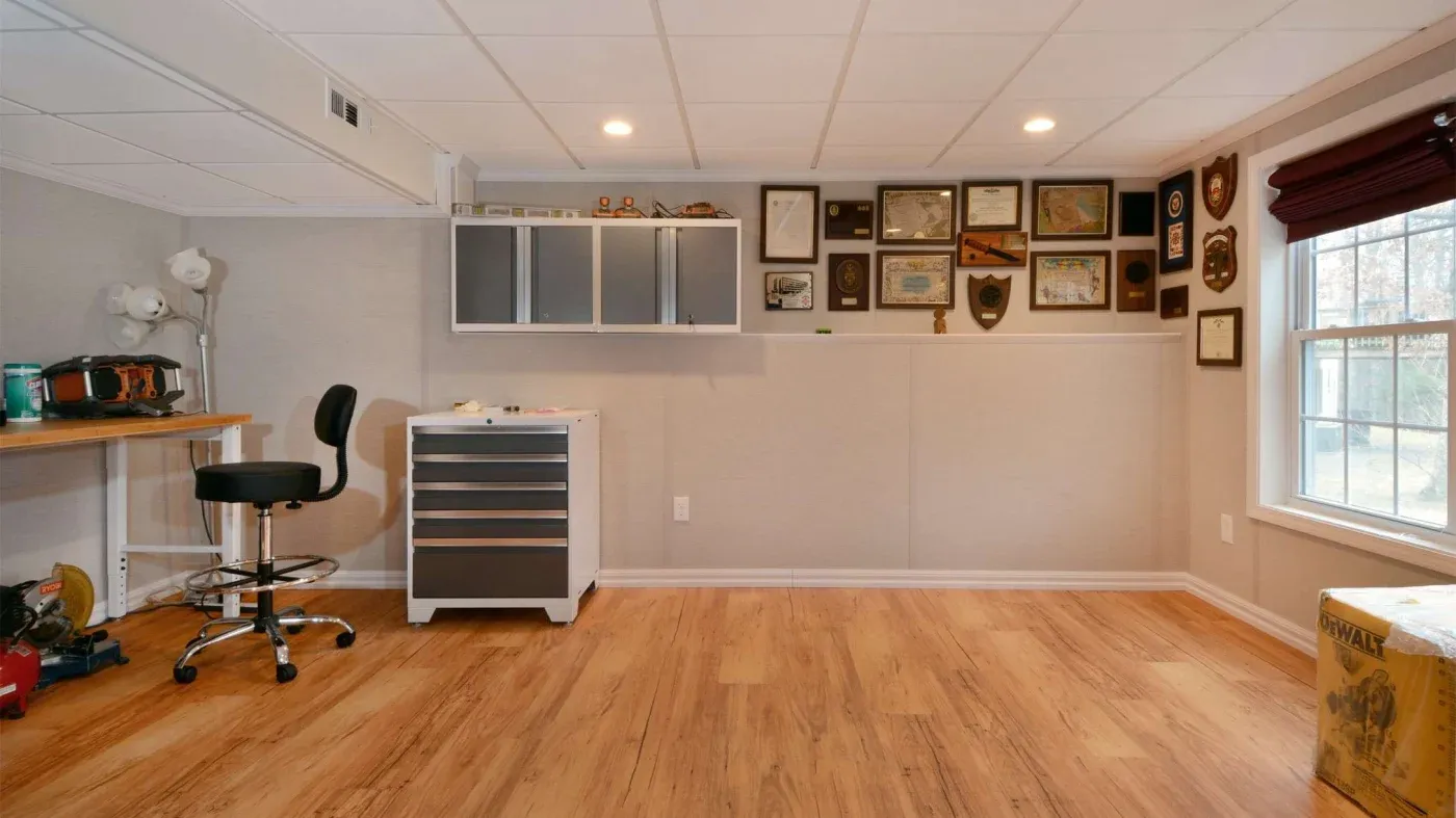 A tidy room with wooden floors, featuring a workbench and swivel chair on the left. The wall displays numerous framed certificates. A window on the right lets in natural light.