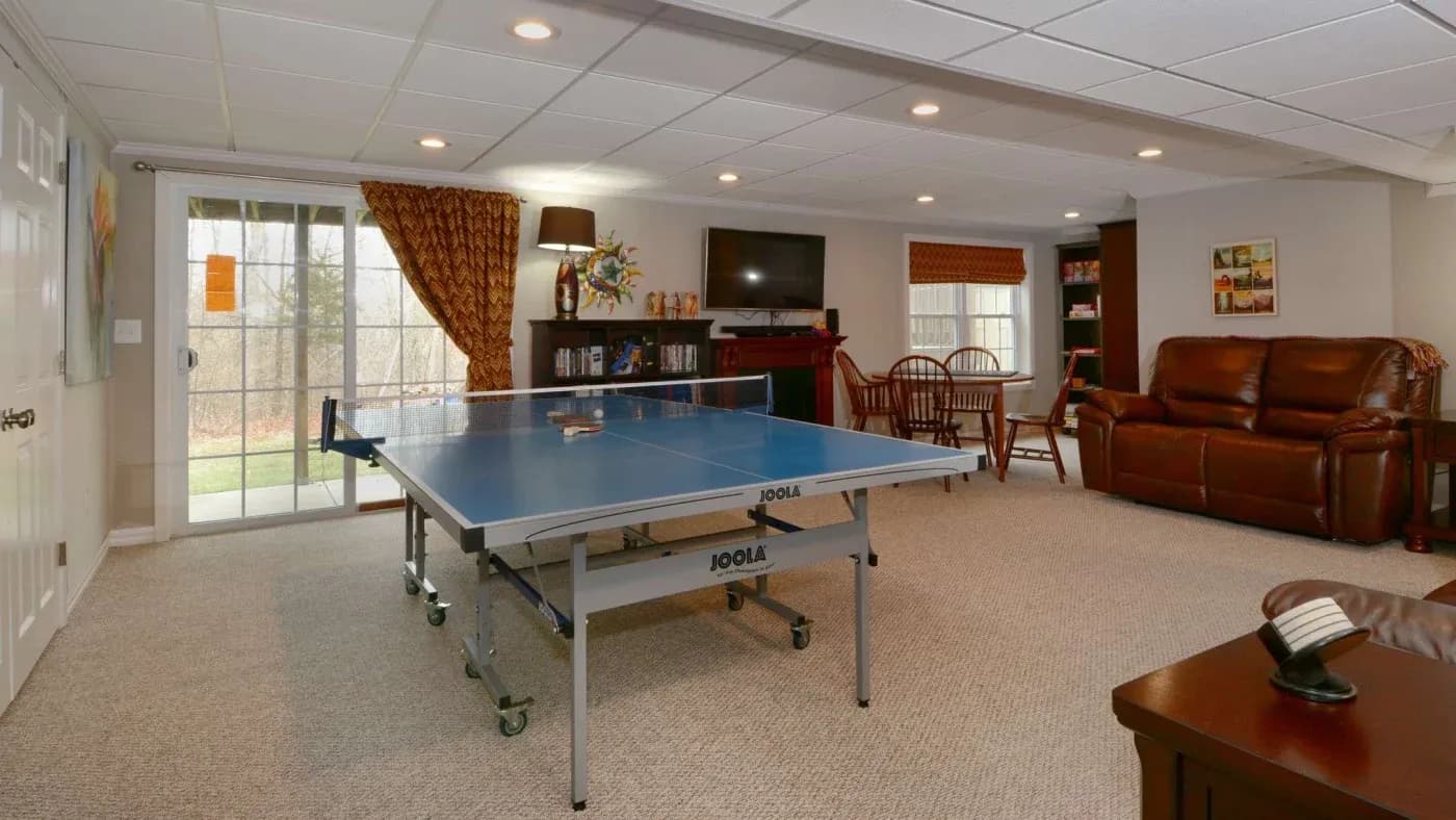 Spacious basement game room with a ping pong table in the center. Plush brown leather sofas, TV, bookshelf, and sliding doors leading outside. Cozy ambiance.