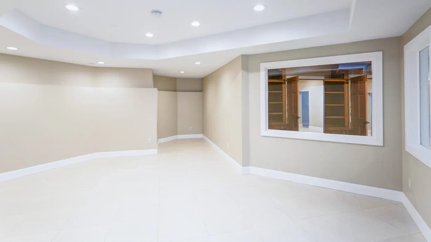 A spacious, neutral-colored room with light beige walls and white trim, featuring recessed ceiling lights and a large window reflecting shelving.