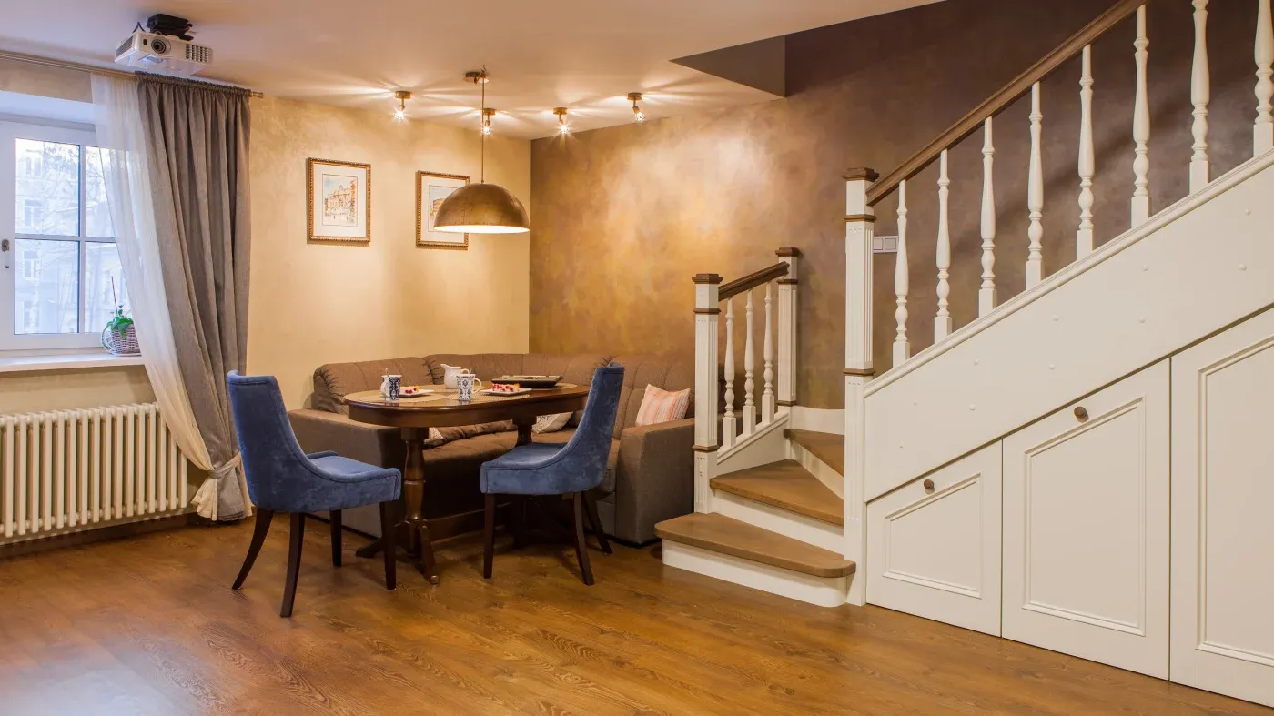 Cozy dining area with a wooden table, blue chairs, and a corner sofa. Warm lighting, soft brown walls, and a staircase with storage beneath.
