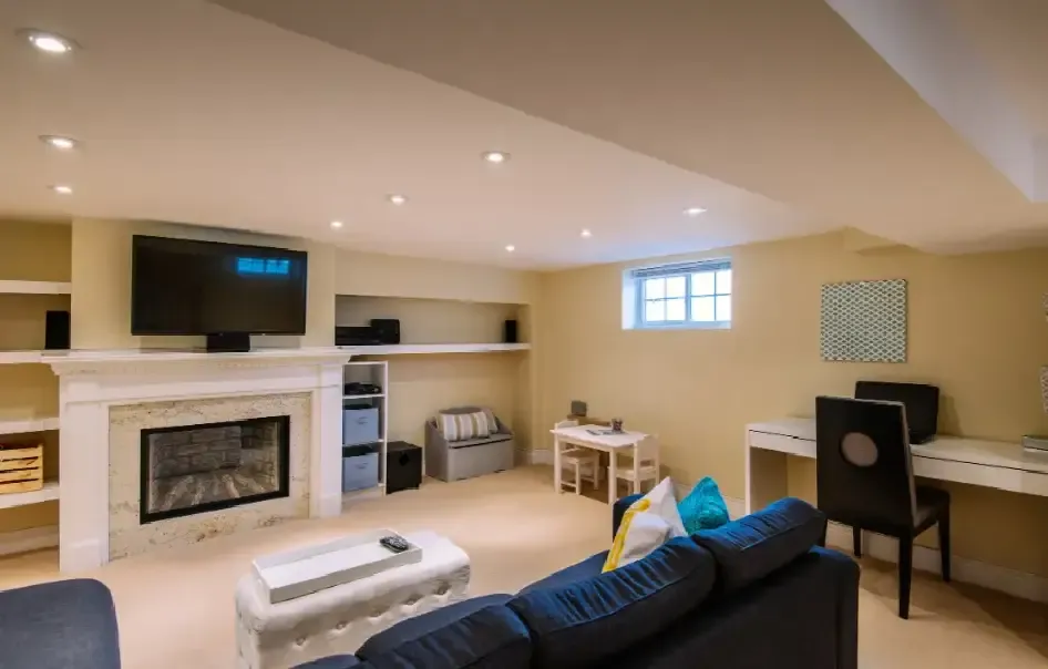 Spacious basement living room with beige walls, recessed lighting, and a cozy vibe. Features include a fireplace, TV, sofa, desk, and shelves.