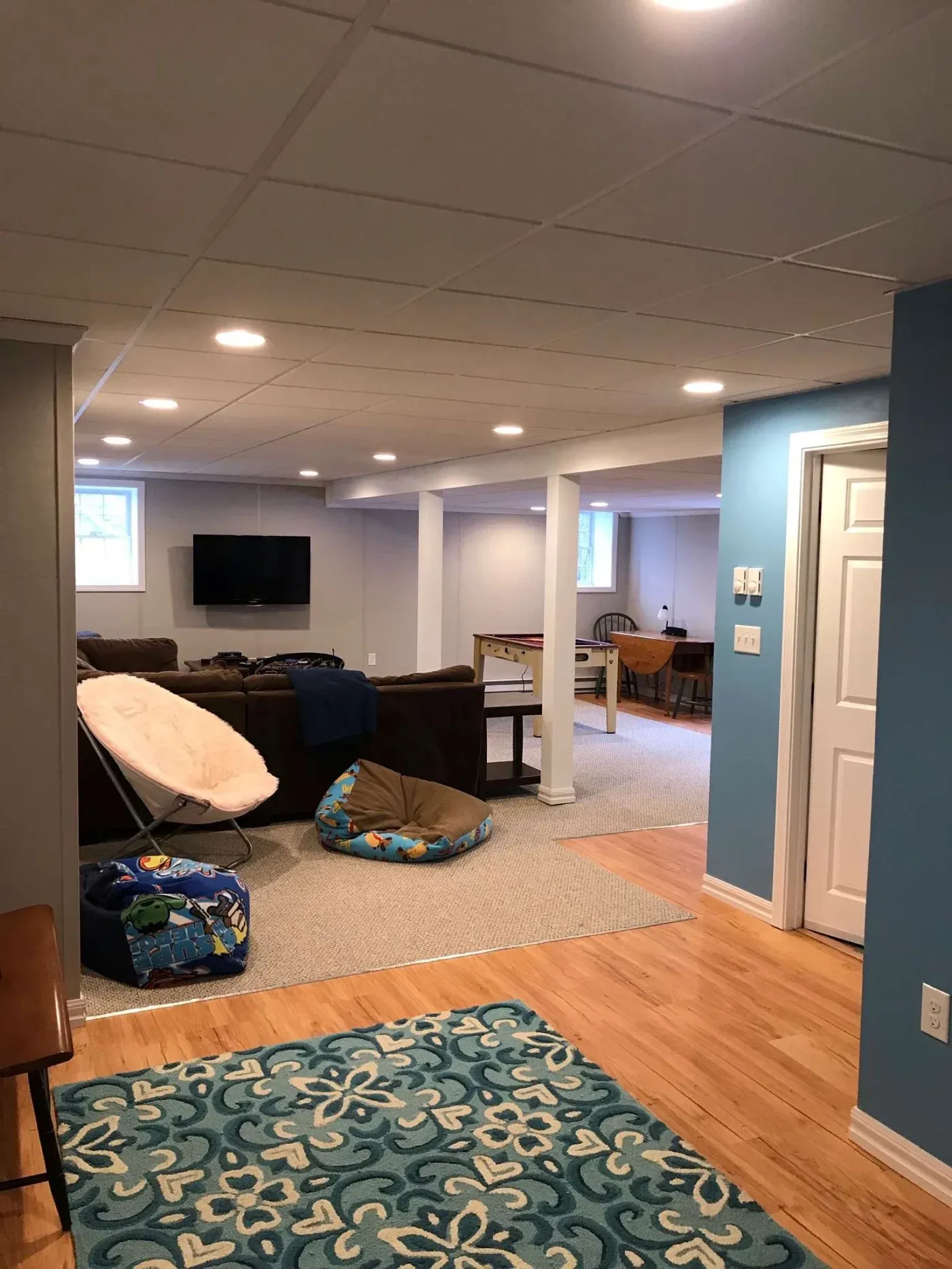 A cozy basement with blue walls and wood flooring features a patterned rug, bean bag, and soft chairs. A TV is mounted on the wall, creating a relaxed atmosphere.