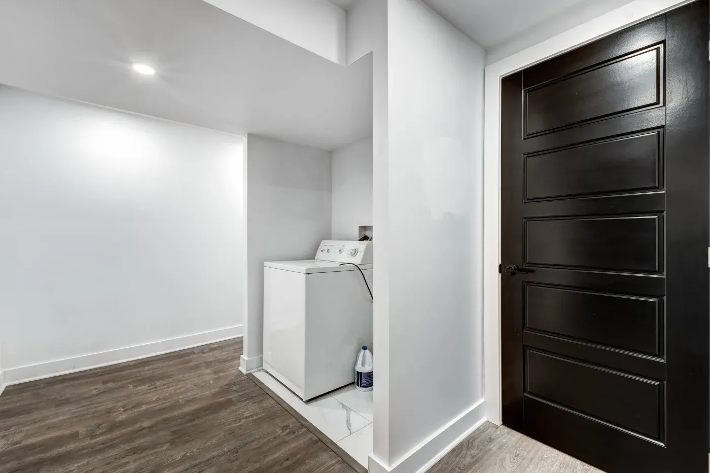 Minimalist laundry area with washer in a white alcove, wood laminate flooring, and a sleek black door. Clean, modern ambiance with bright lighting.